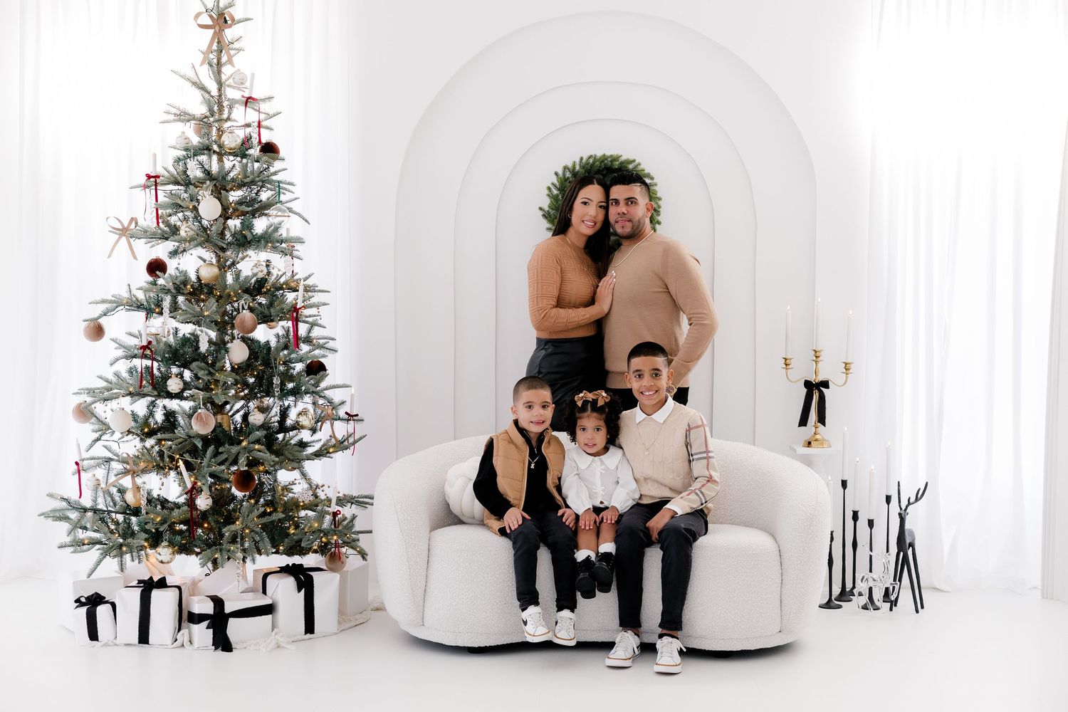 A family poses together on a white sofa with decorated Christmas trees in an elegant studio setting.