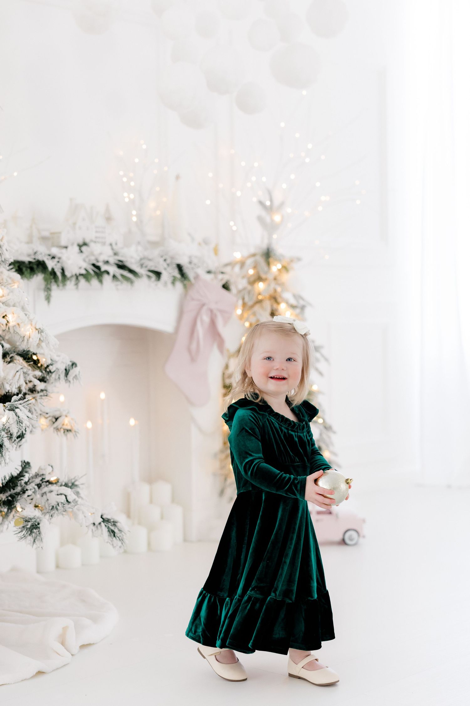 Baby wearing emerald green velvet dress playing near white Christmas decorations and lights.