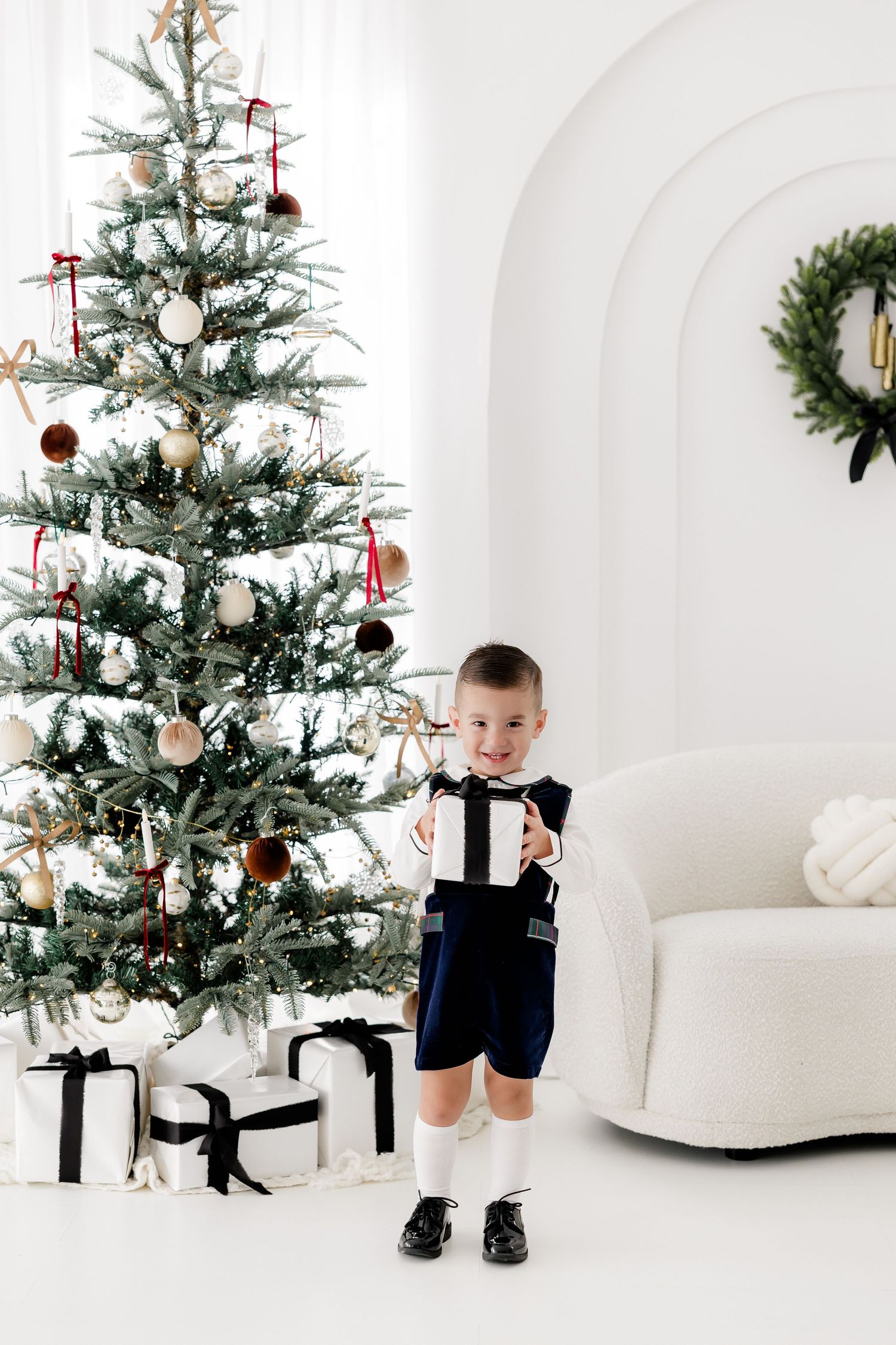 Small child in formal wear standing by decorated Christmas tree with wrapped presents.