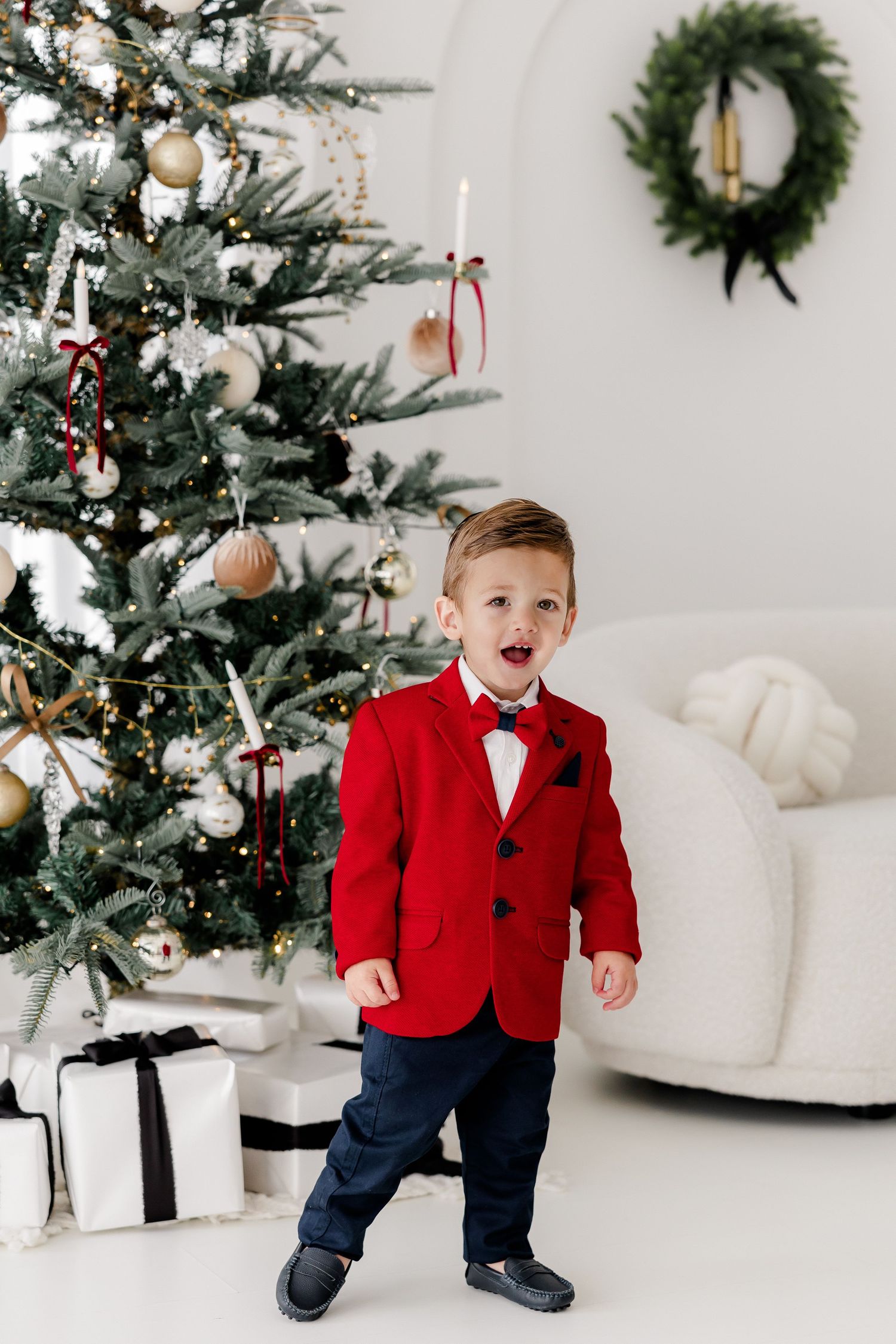 Child in red blazer and navy pants poses energetically by decorated Christmas tree.
