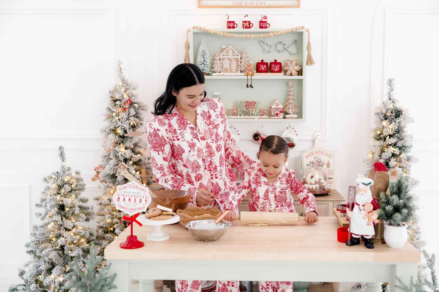 Mother and daughter in matching red floral Christmas pajamas baking together in a festive holiday kitchen with decorated trees.