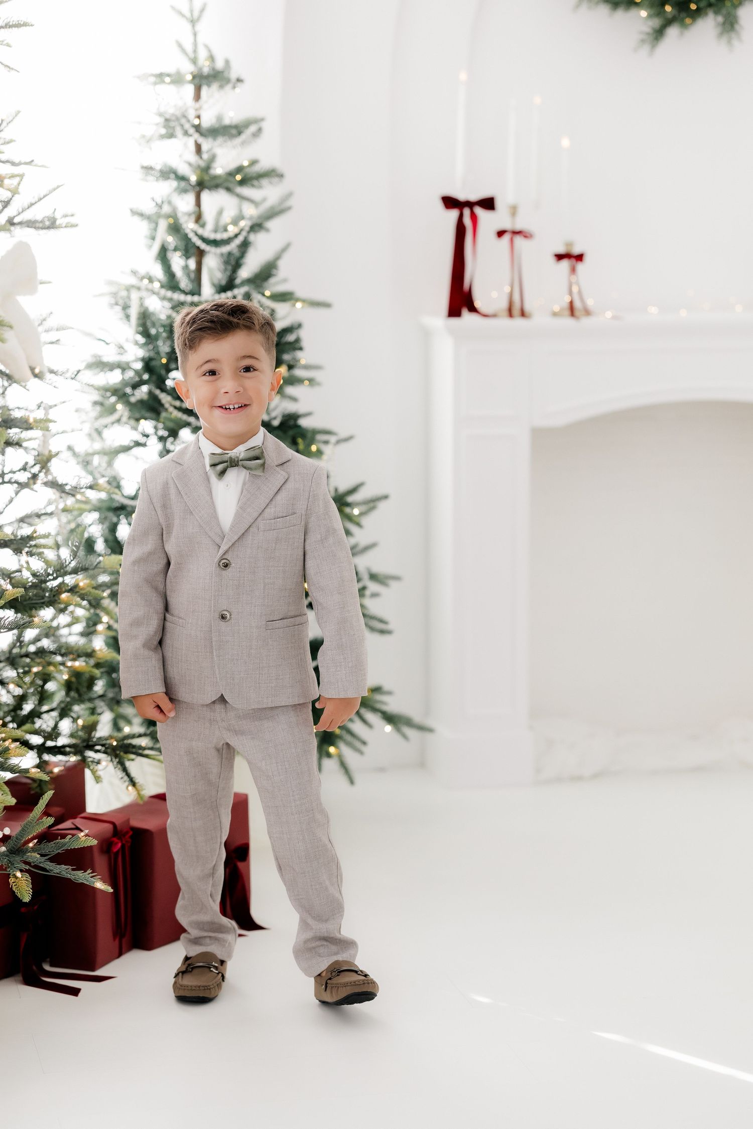 Young boy in gray suit with bow tie smiling by Christmas tree and fireplace with red gifts.