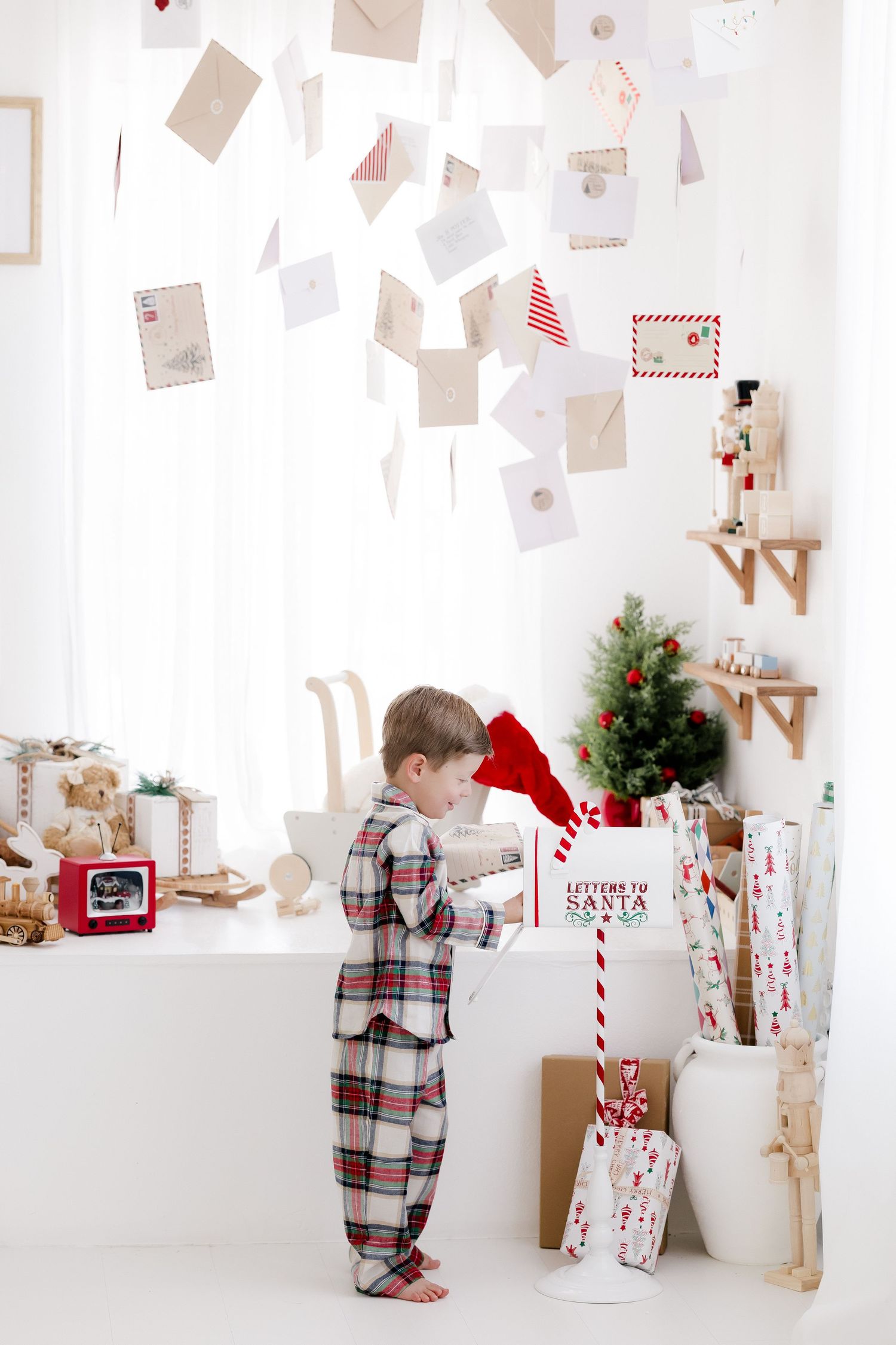 Child in plaid pajamas writing a letter to Santa Claus beside a festive mailbox in a decorated Christmas room.