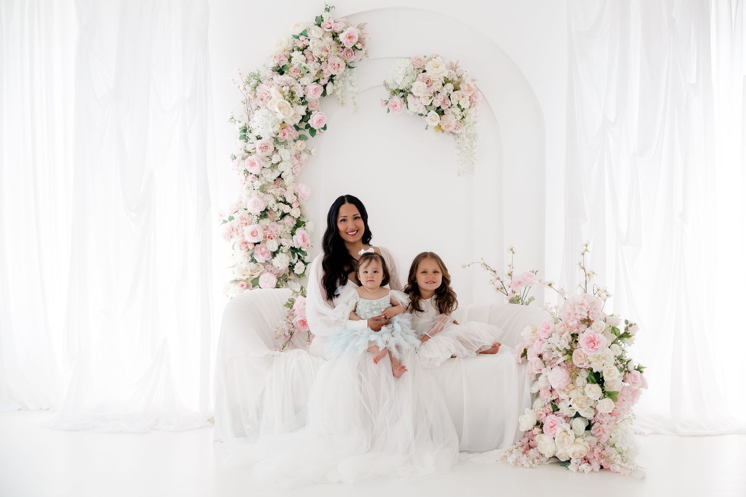 A white floral arch with pink and blush roses creates an elegant backdrop for a wedding photo session.