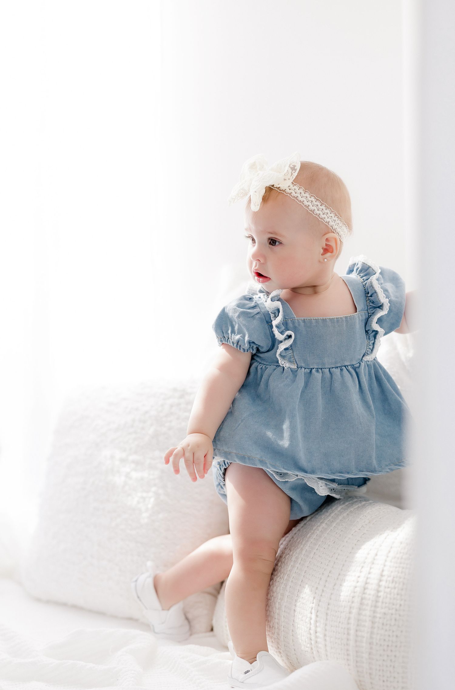 Baby in light blue ruffled dress sits on white textured cushion while looking to the side.