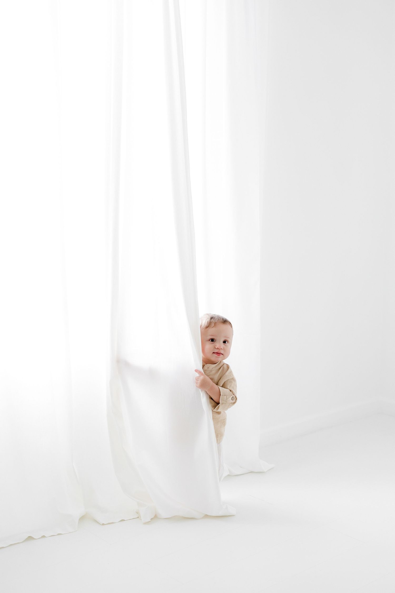 A baby peeks playfully from behind white curtains in a bright, minimalist setting.