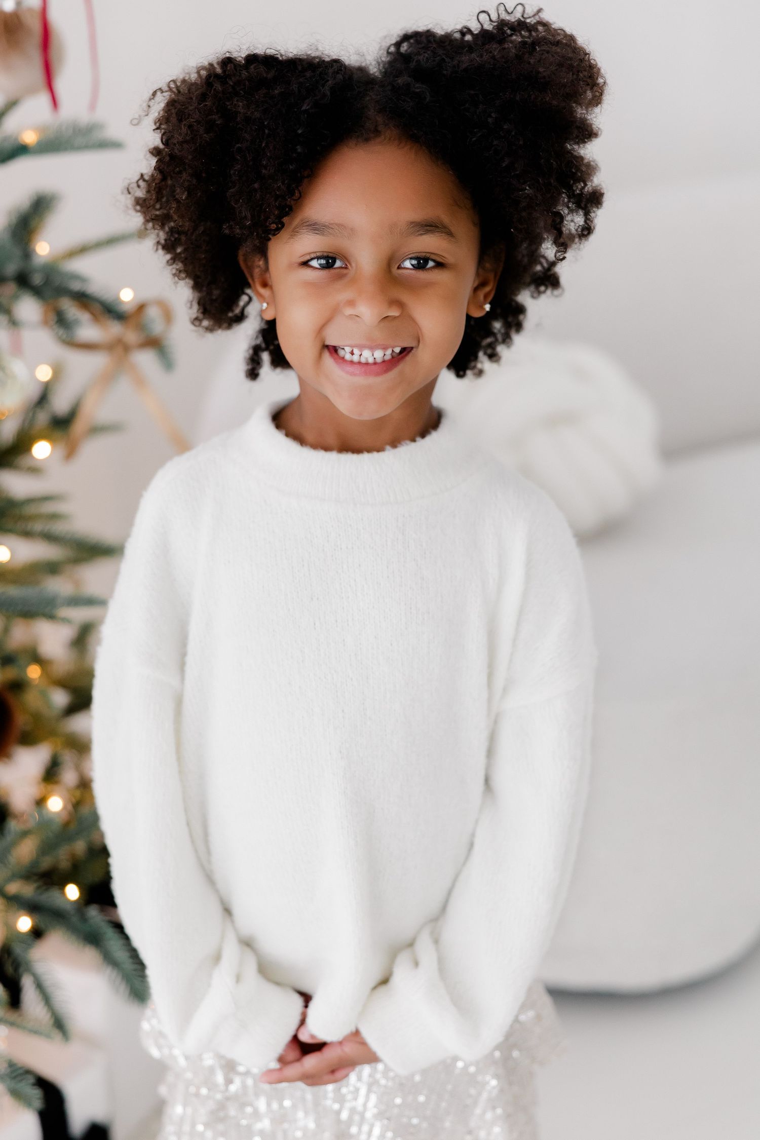 Child in white sweater poses in front of Christmas tree in bright studio setting.