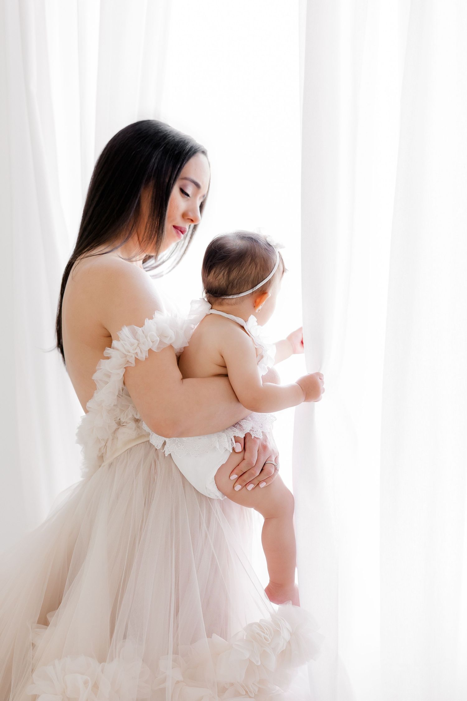 A tender moment captured in soft light as a parent in white lace holds their baby against sheer curtains.
