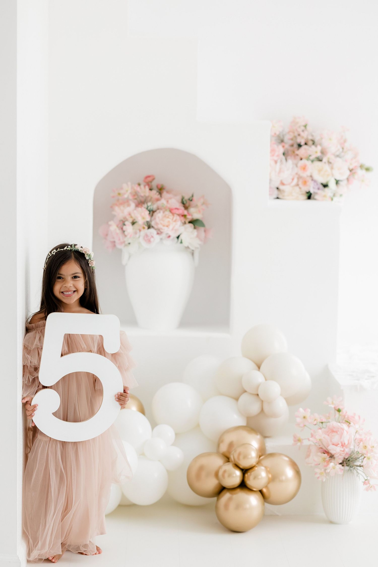 Elegant birthday setup with large number 5, pink tulle dress, white and gold balloons, and delicate floral arrangements.