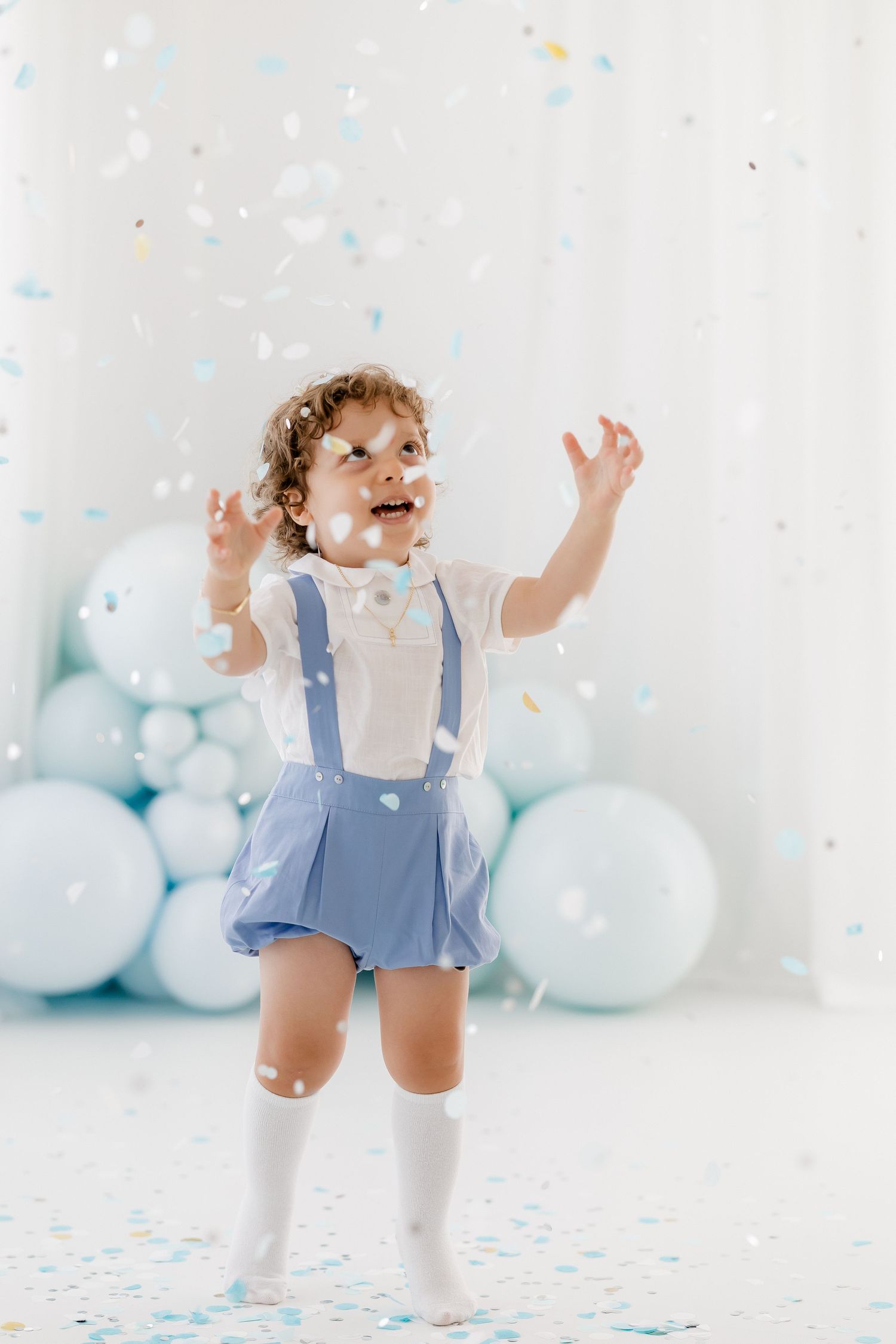 Toddler in blue overalls and white socks dances joyfully among pastel blue balloons and confetti.