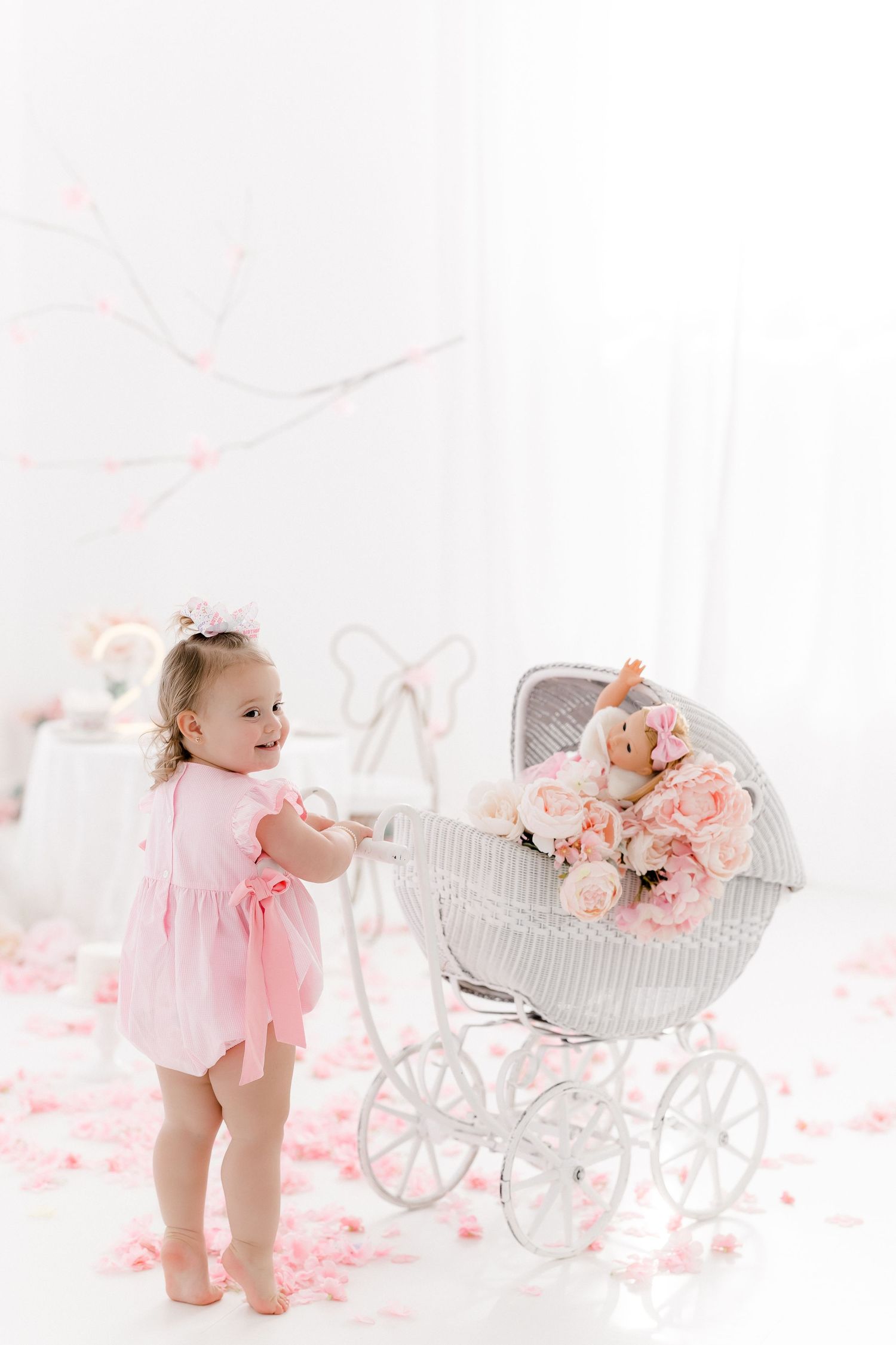 Baby in pink dress plays near vintage carriage with floral decorations in dreamy white studio setting.