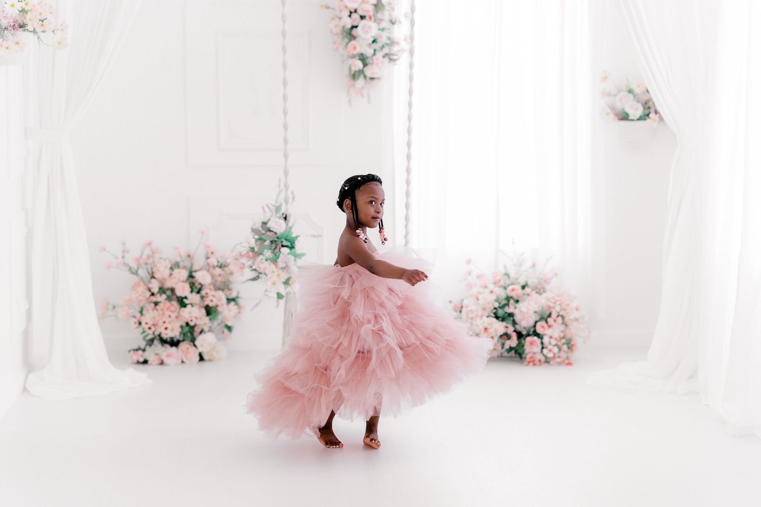A child in a pink tulle dress twirls gracefully in an elegant white room decorated with delicate floral arrangements.