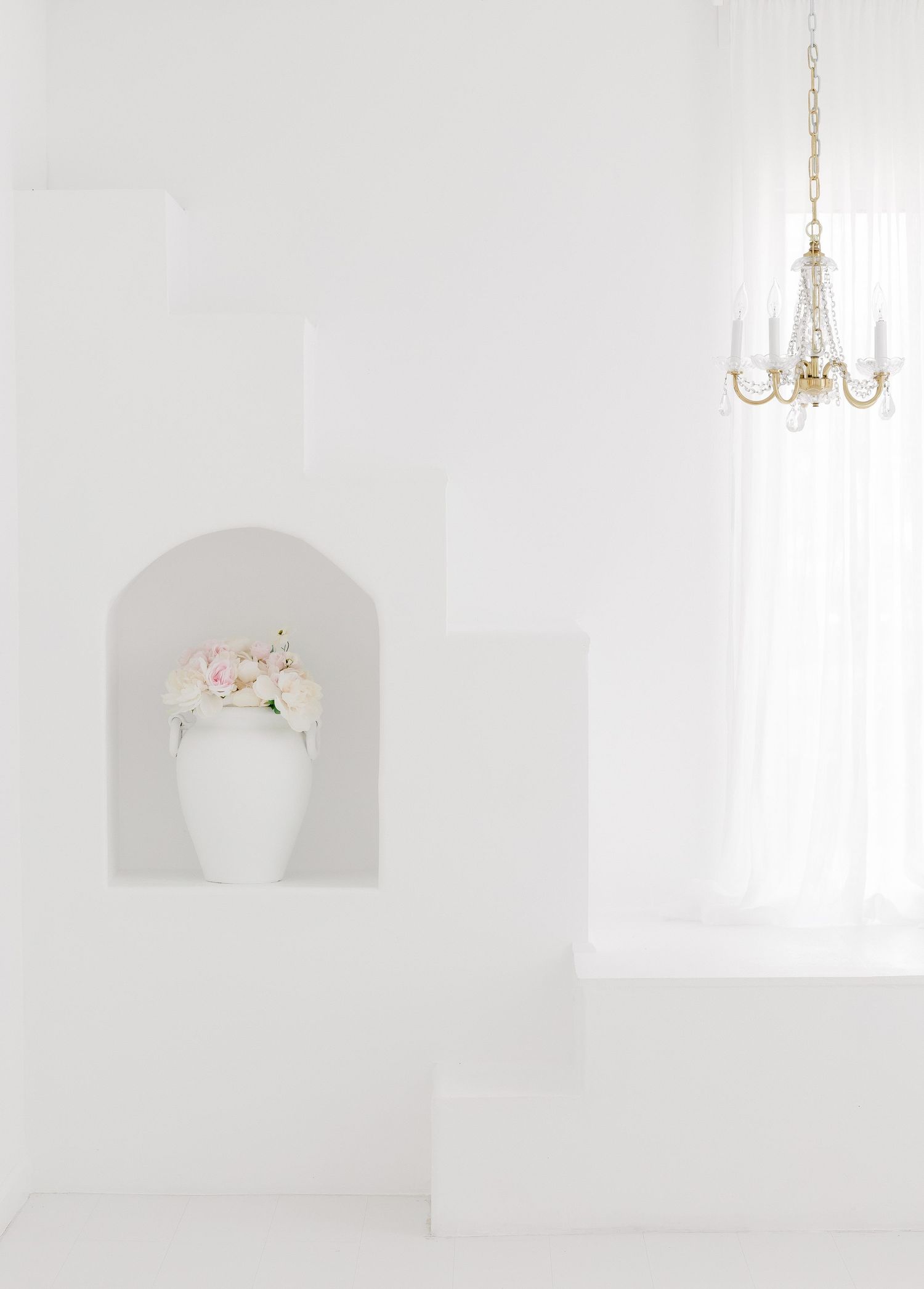 A serene white interior with chandelier and arched wall niches showcases elegant minimalist design elements.