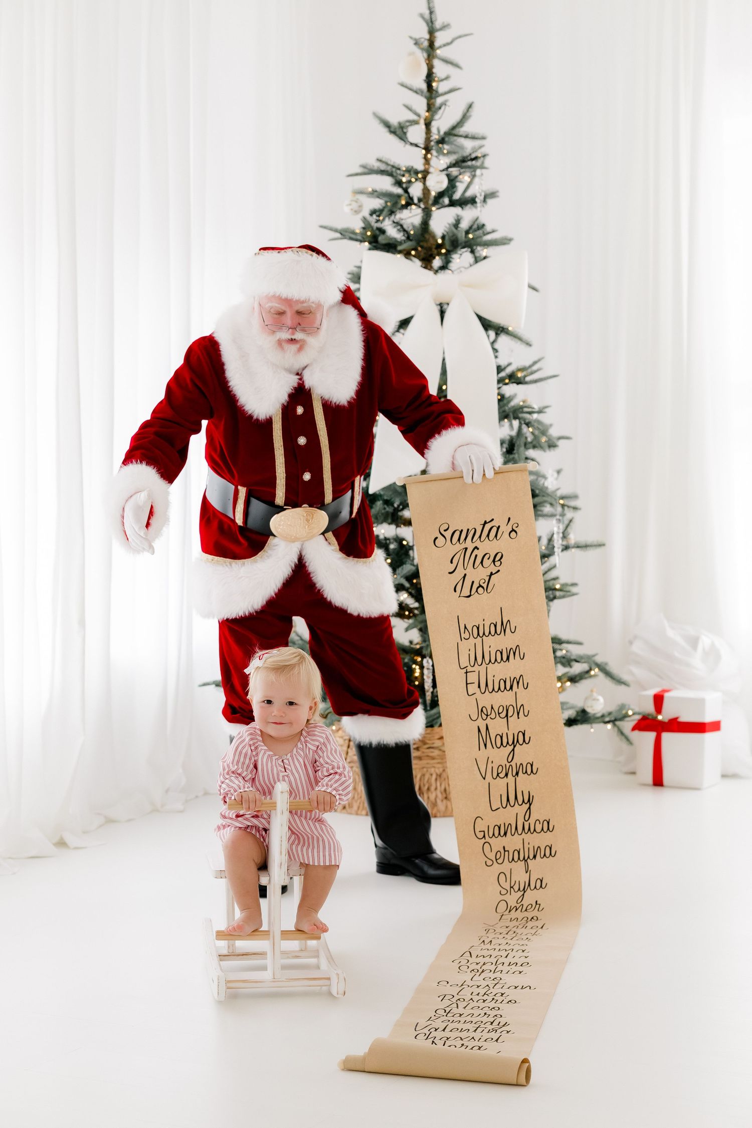 Santa Claus holds his Nice List scroll beside a toddler on a rocking horse near a decorated Christmas tree.