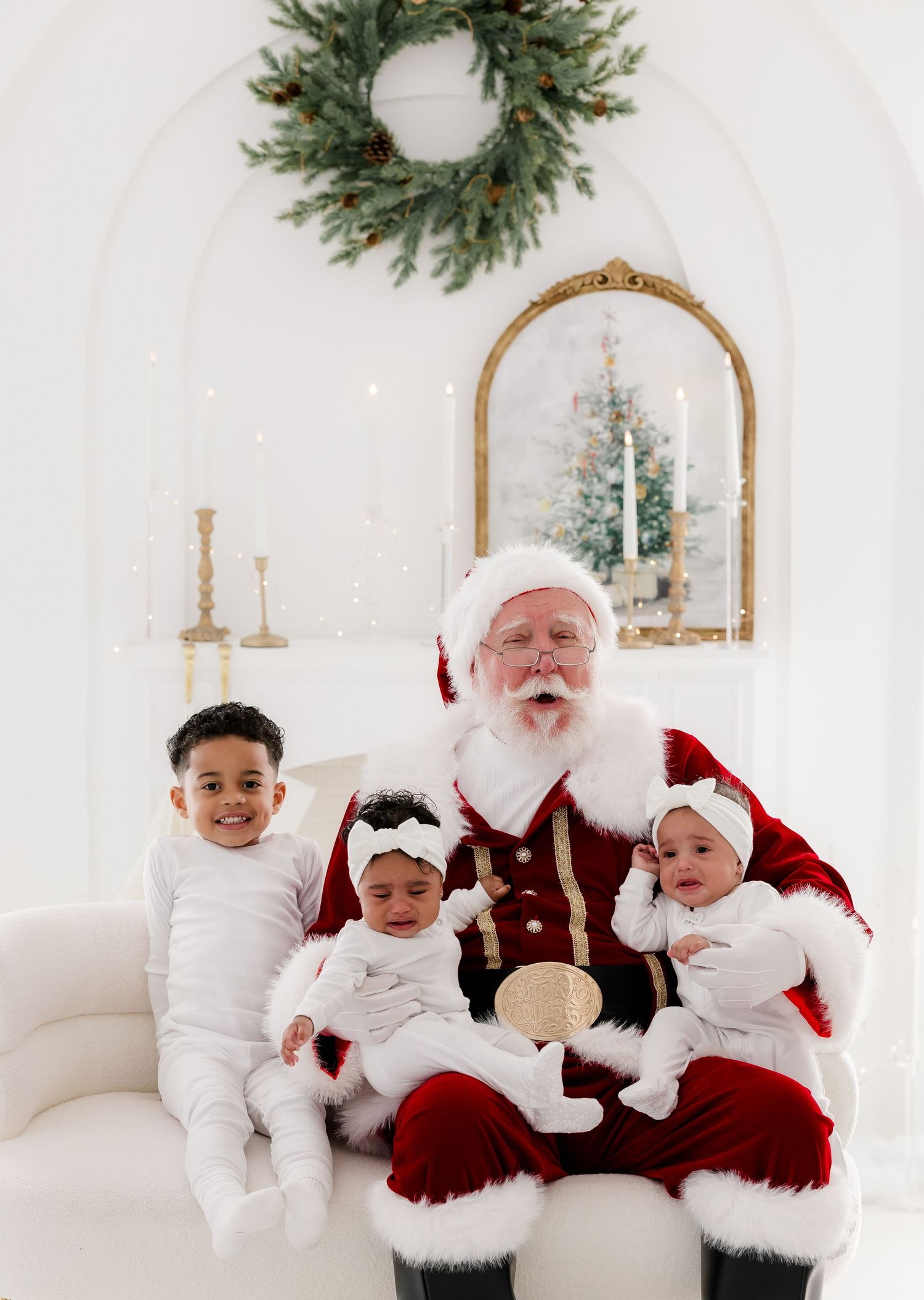 Three young children sit with Santa Claus on a white sofa in a bright, elegant Christmas photo studio.