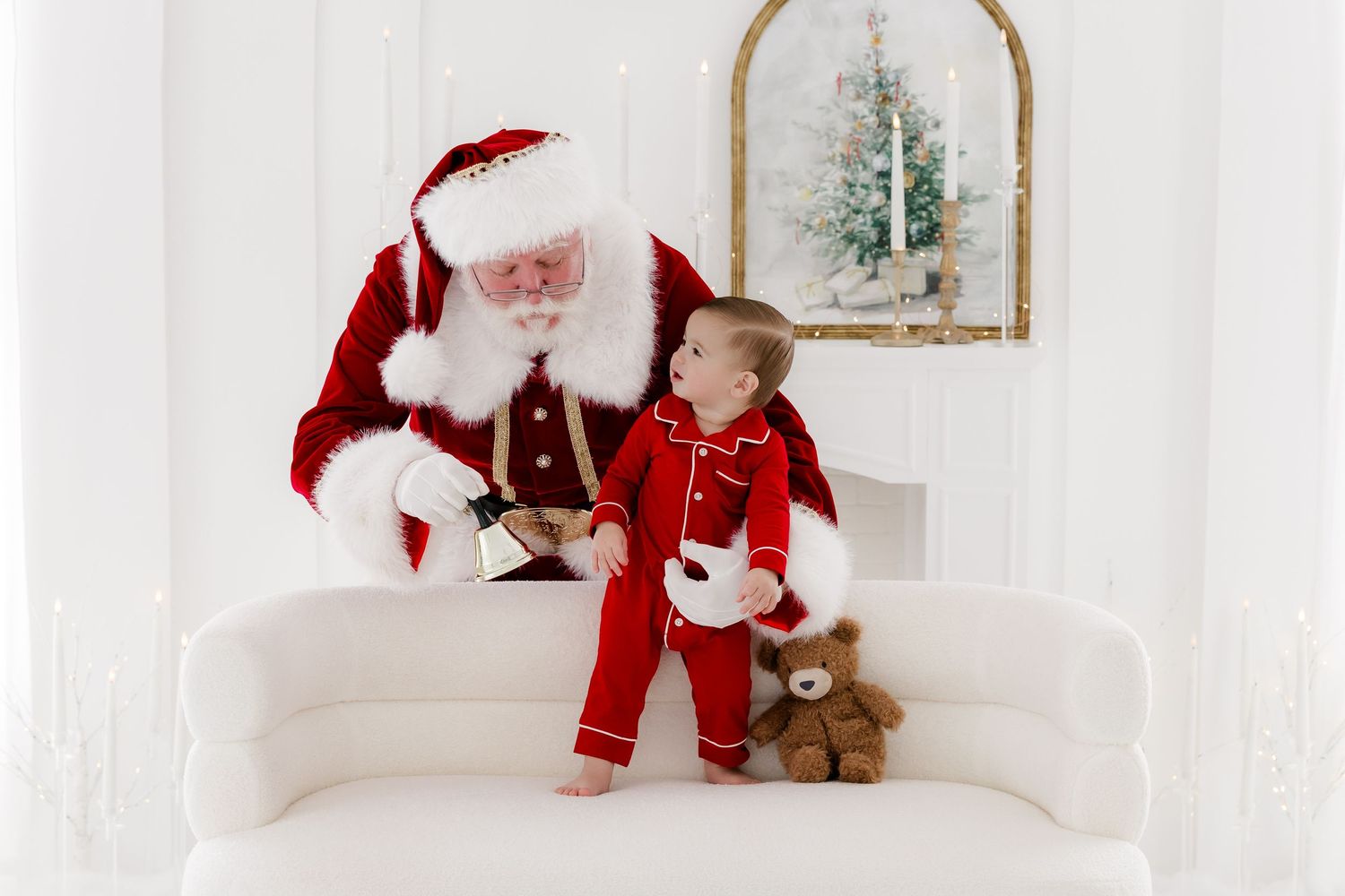 Santa Claus in red suit sits on white sofa with toddler in red pajamas beside teddy bear in bright white holiday studio.