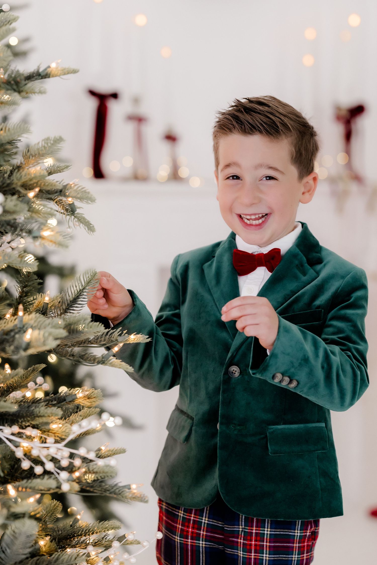 A cheerful boy in a green velvet blazer and red bow tie smiles beside a decorated Christmas tree.