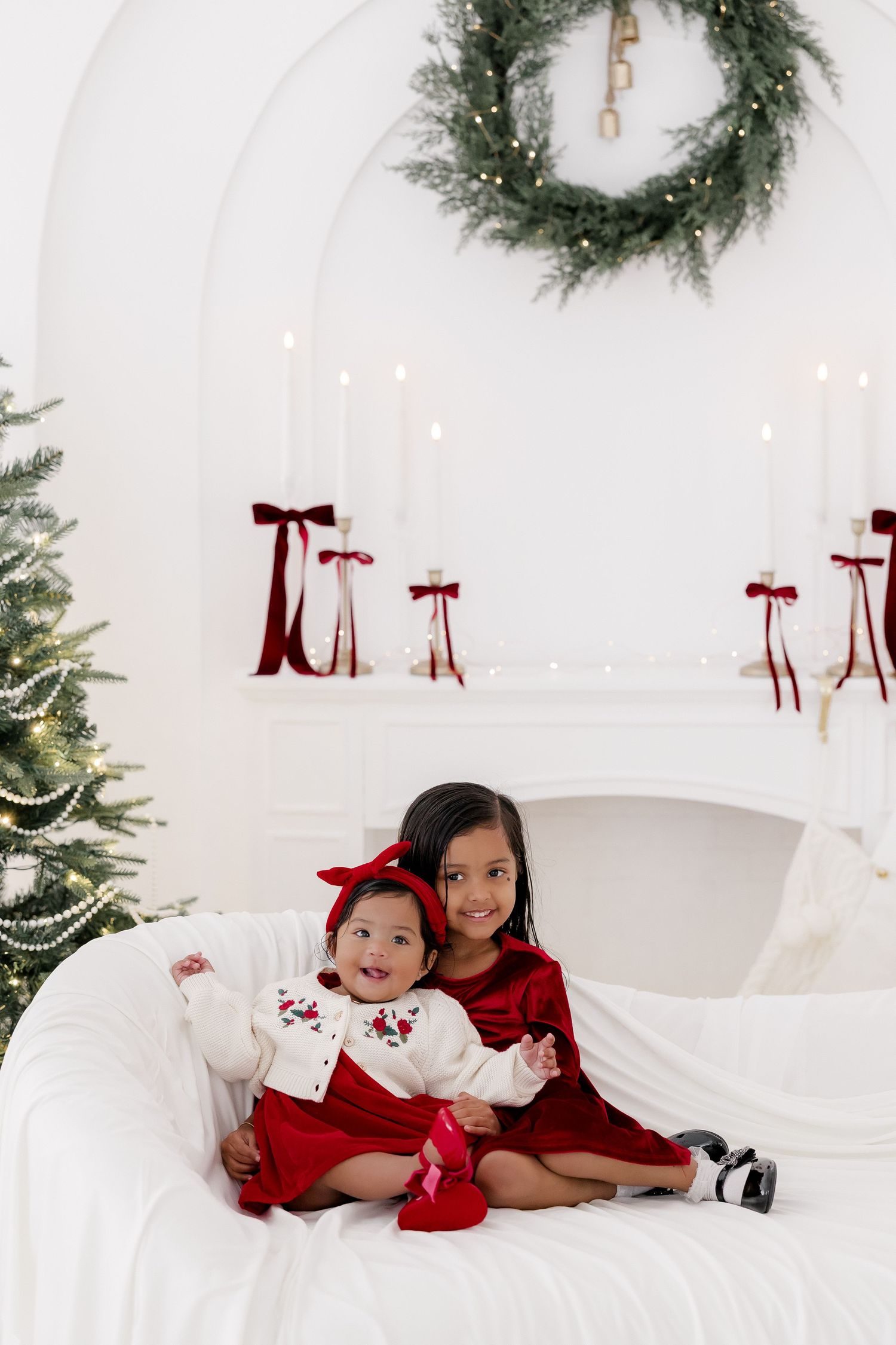 Two young sisters in red holiday dresses pose together on a white sofa with Christmas decorations behind them.