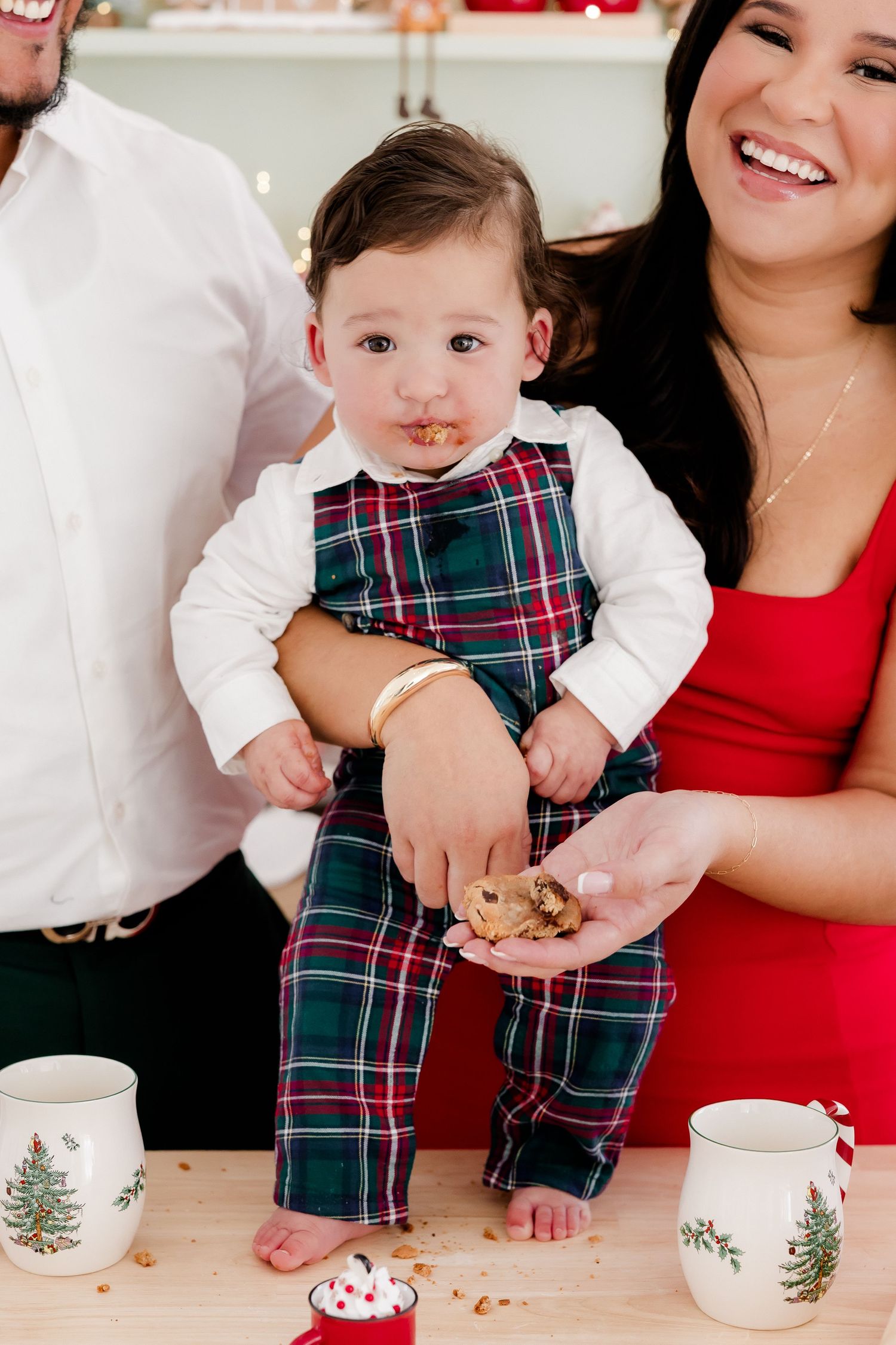A baby in plaid Christmas overalls eats a cookie while being held by laughing parents near holiday mugs.