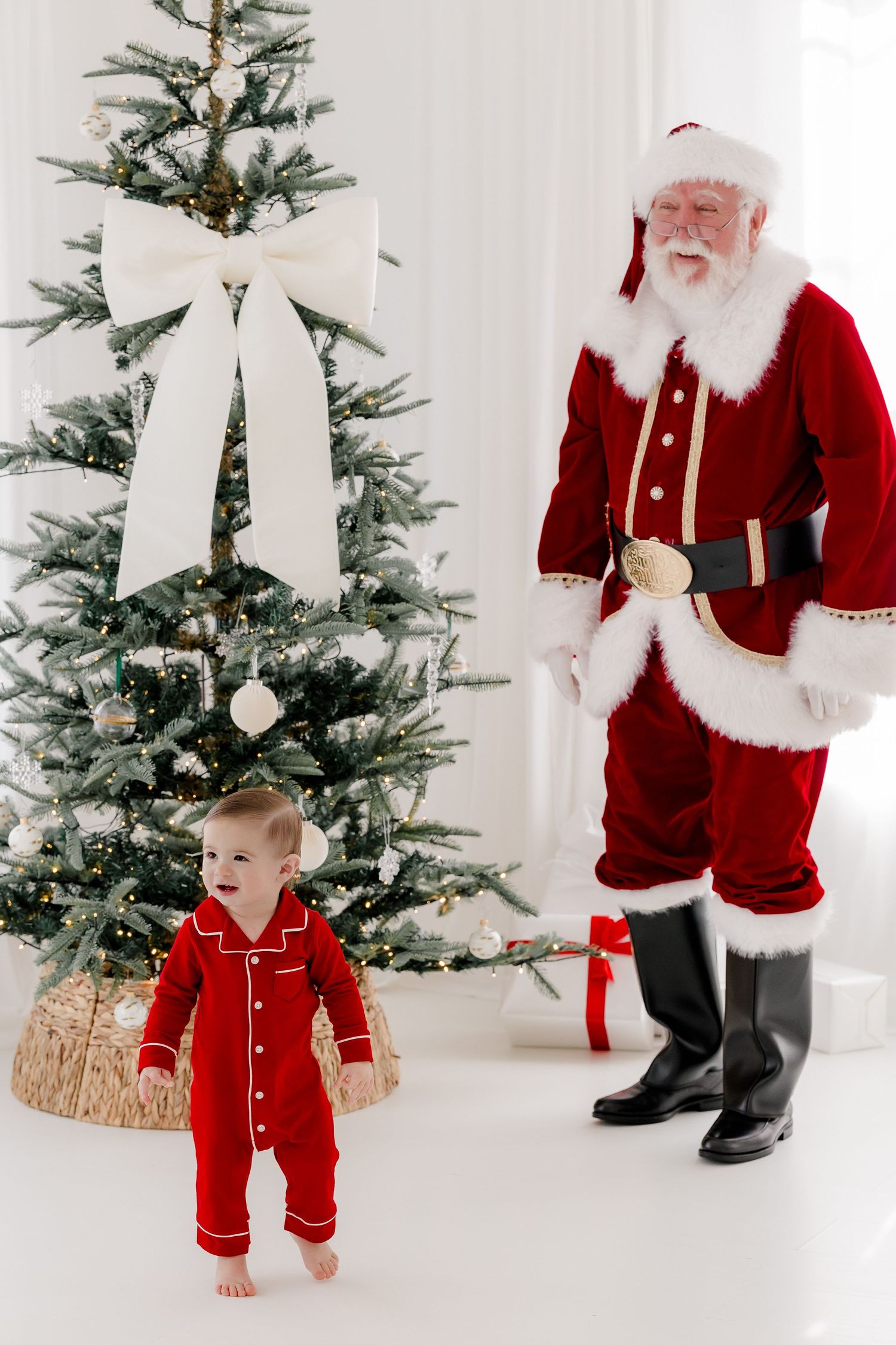 Toddler in red pajamas stands by decorated Christmas tree next to Santa Claus indoors.