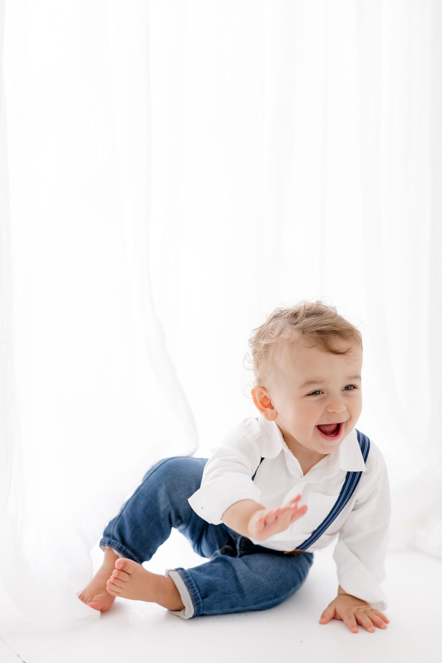 Happy toddler wearing white shirt and blue jeans with suspenders sits on white background while laughing.