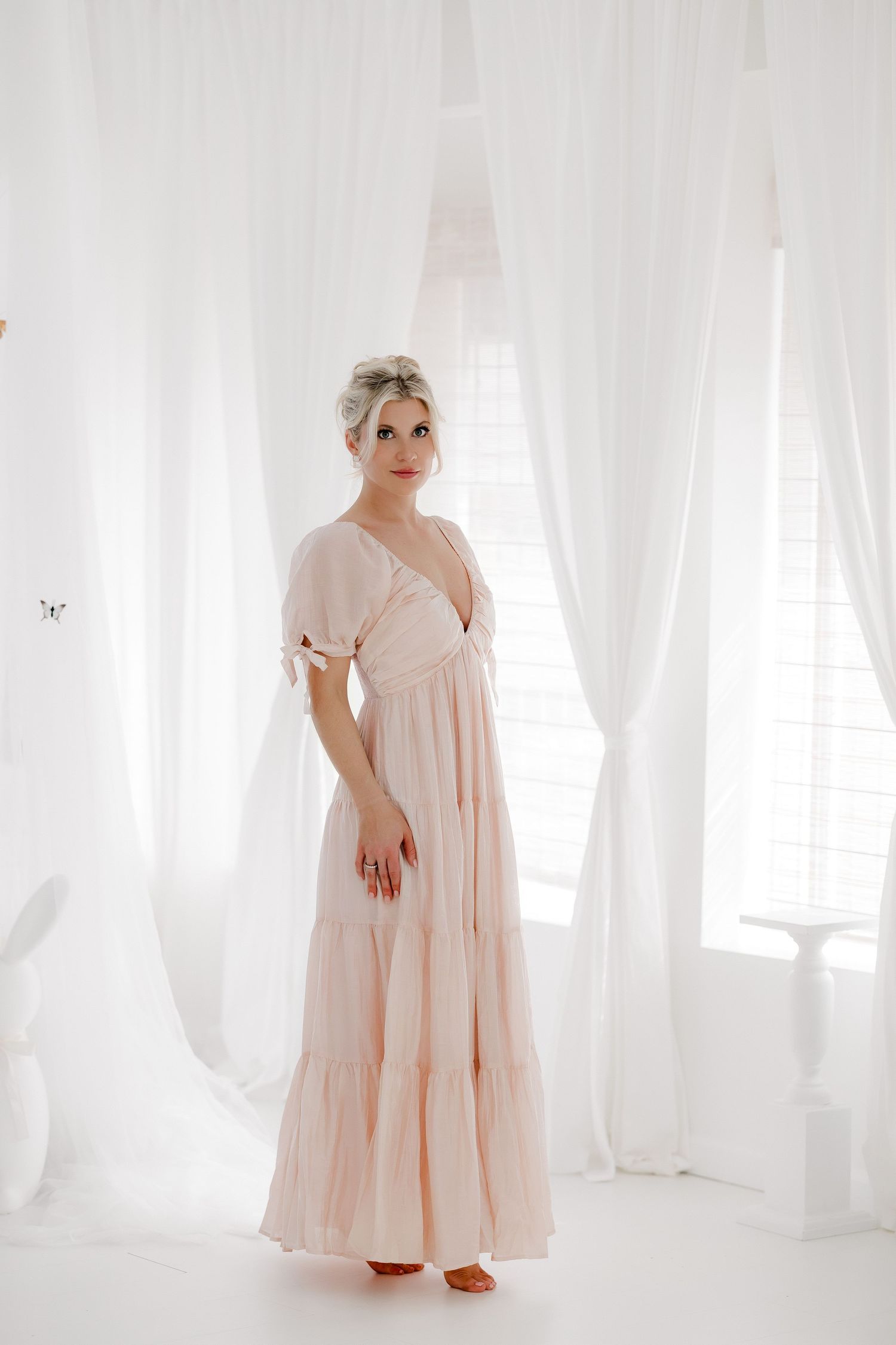 A figure in a flowing blush pink dress stands gracefully in a bright white room with sheer curtains.