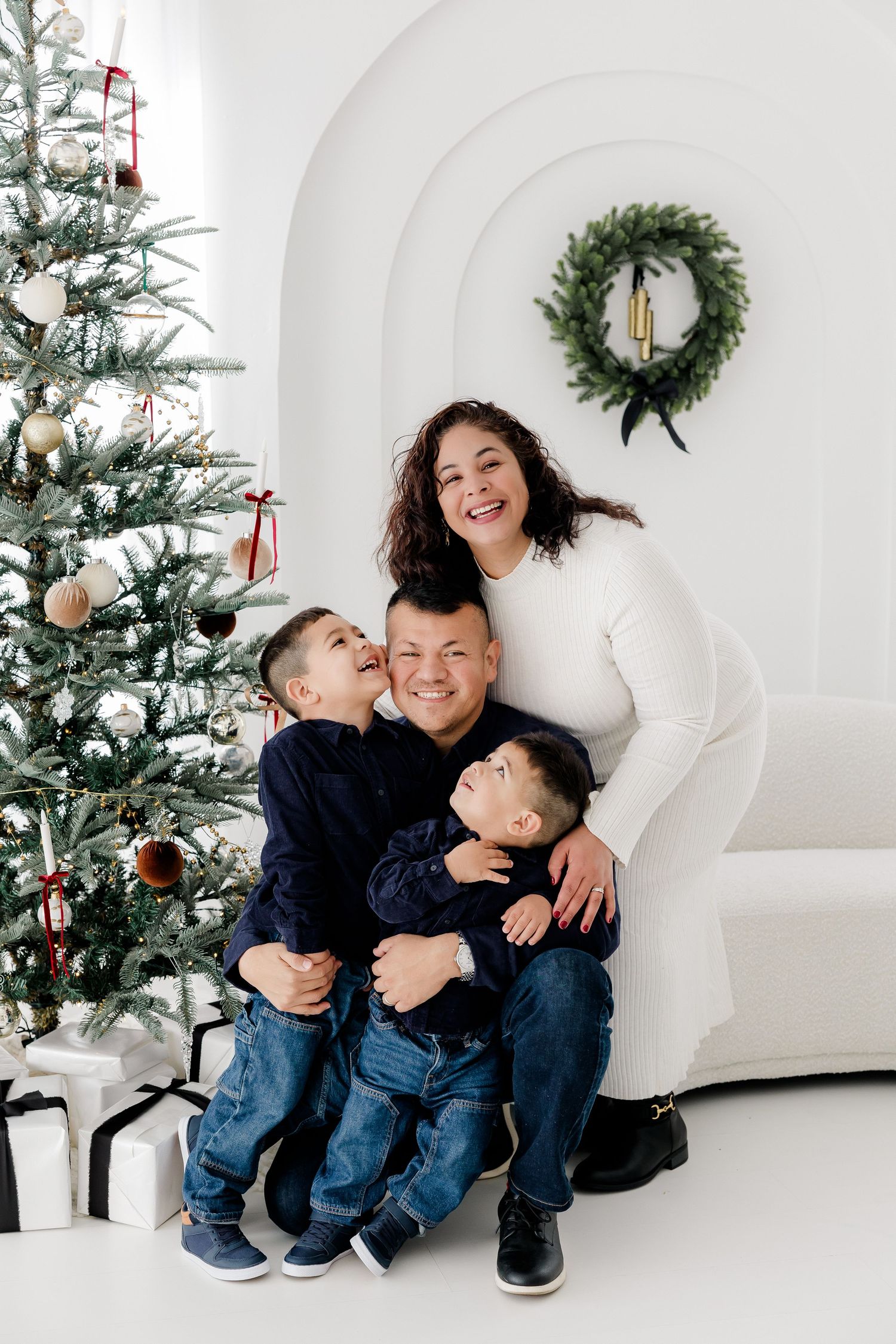 A family shares joyful moments next to a Christmas tree in a bright white studio setting.