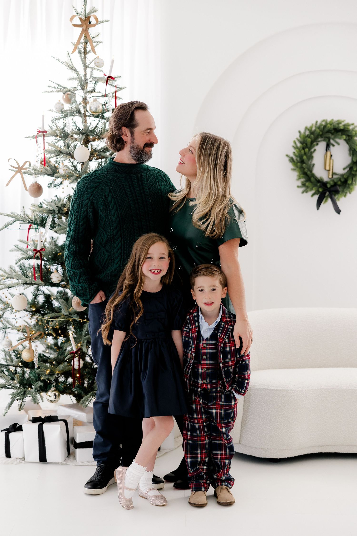 A family poses together in matching outfits near a decorated Christmas tree and wrapped presents in a white studio setting. nj holiday minis