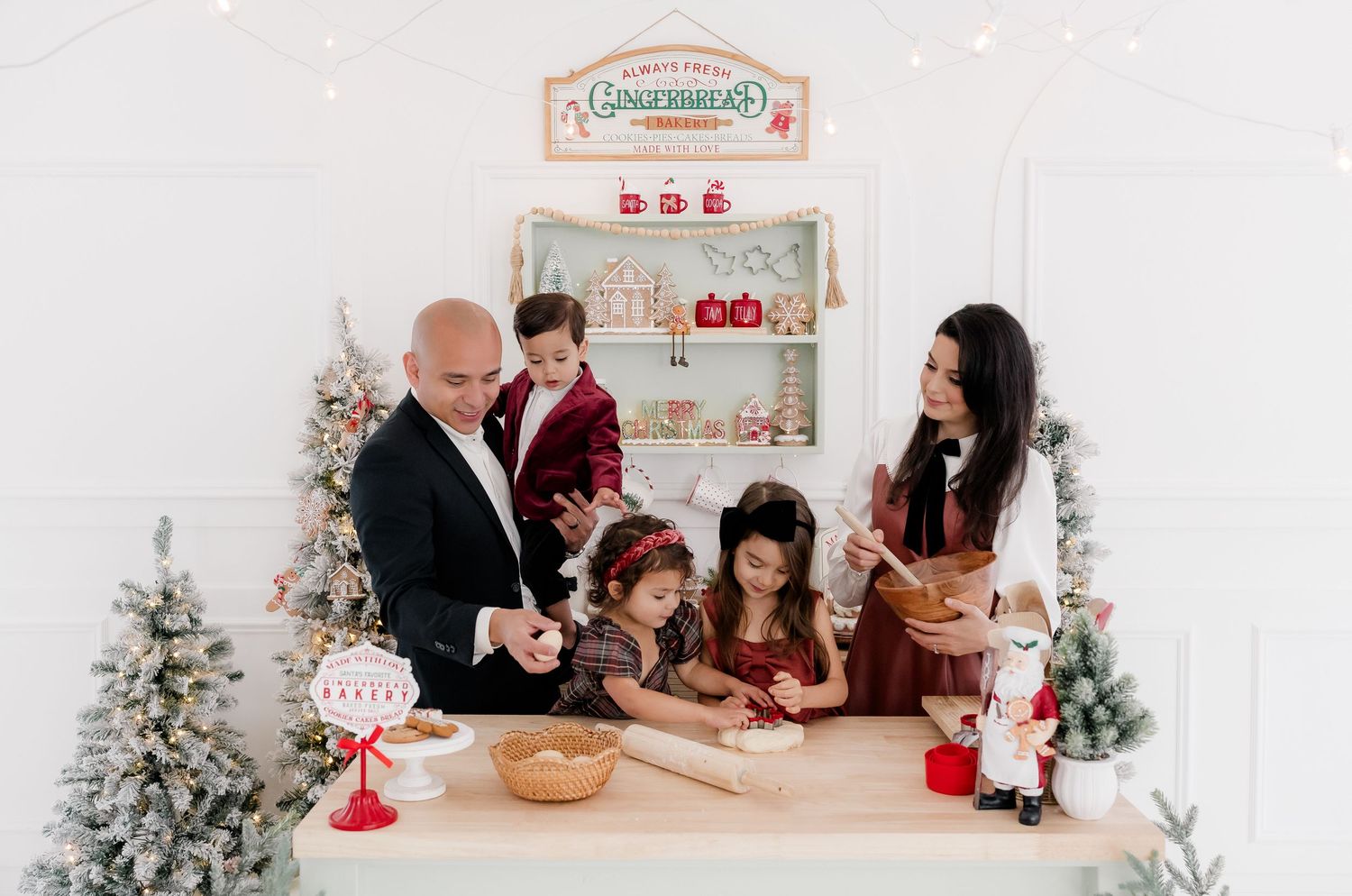 Family of five poses at a festive gingerbread stand with Christmas trees and holiday decorations in a white studio.