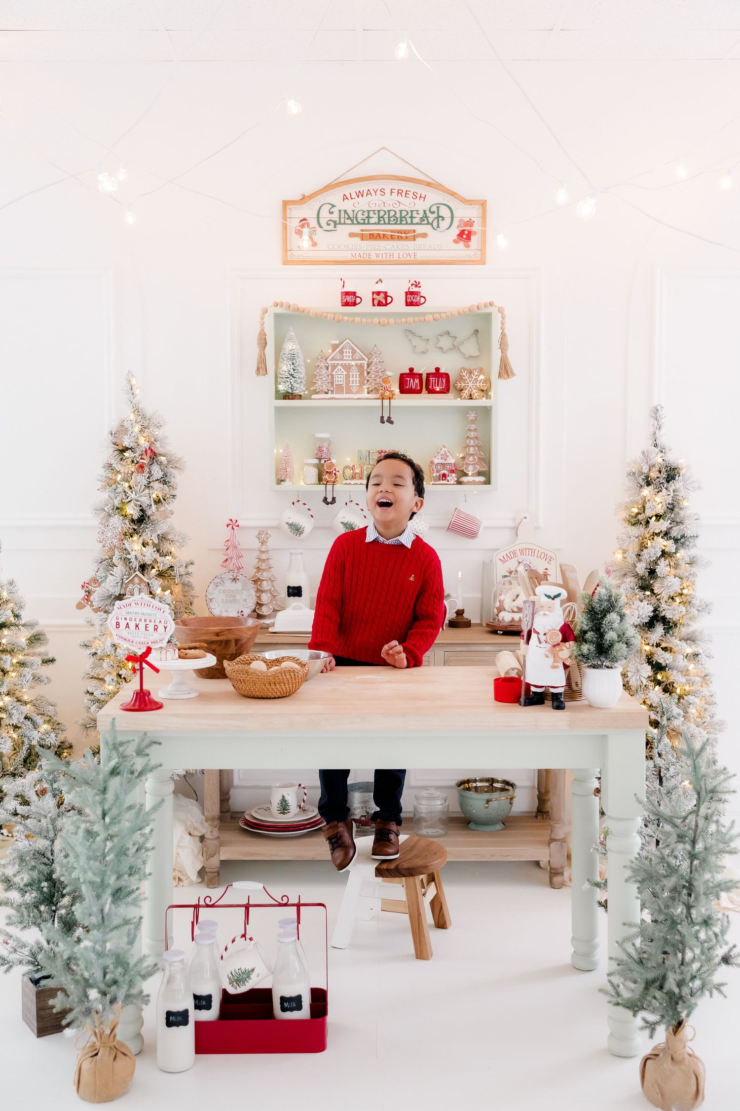 A joyful boy in a red sweater stands at a festive gingerbread bakery table surrounded by snowy Christmas trees and holiday decor.