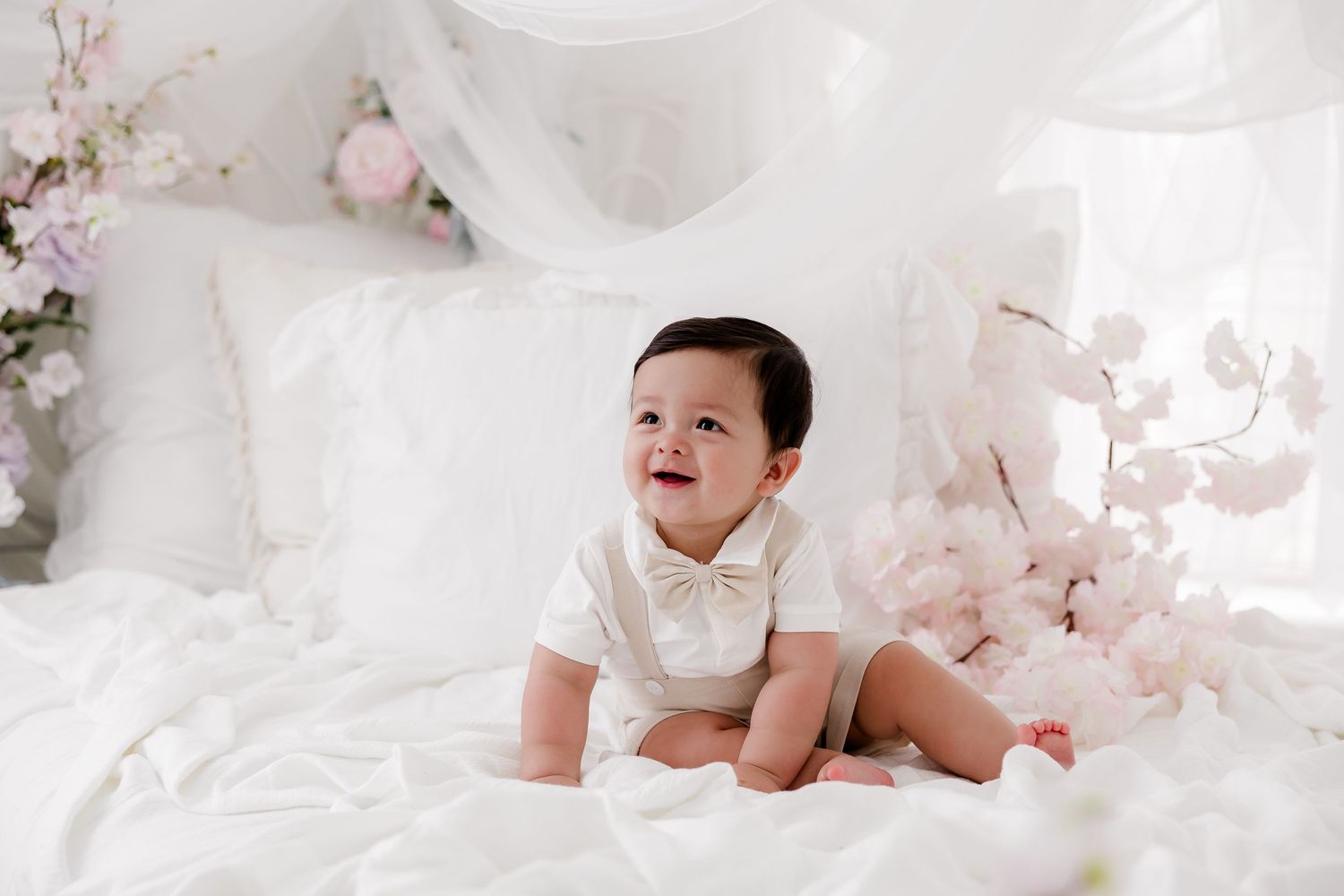 A baby in a white outfit crawls on soft white bedding with pink floral decorations in the background.