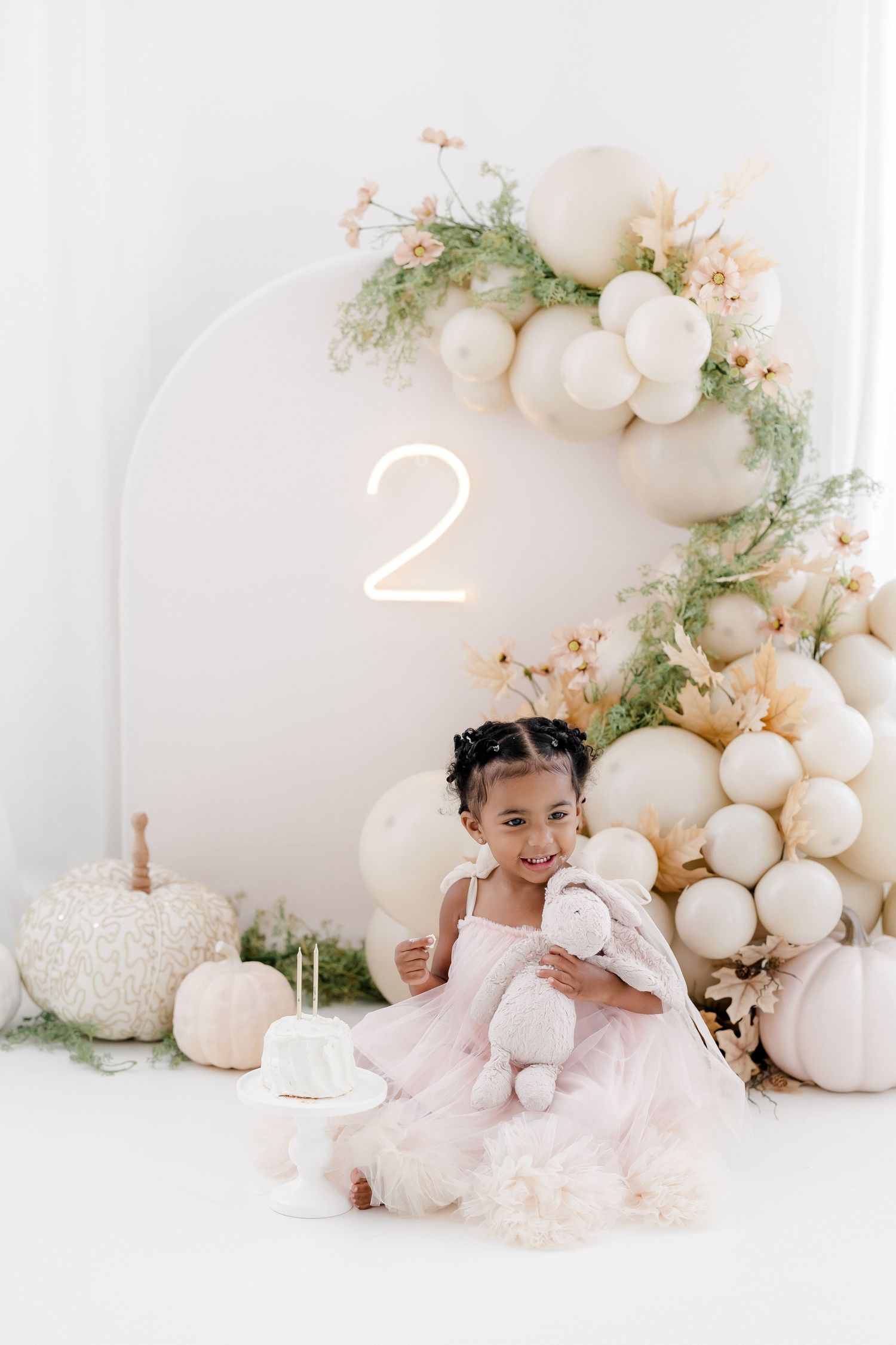 Celebration setup with number 2 light, white pumpkins, and balloon arch decorated with flowers.