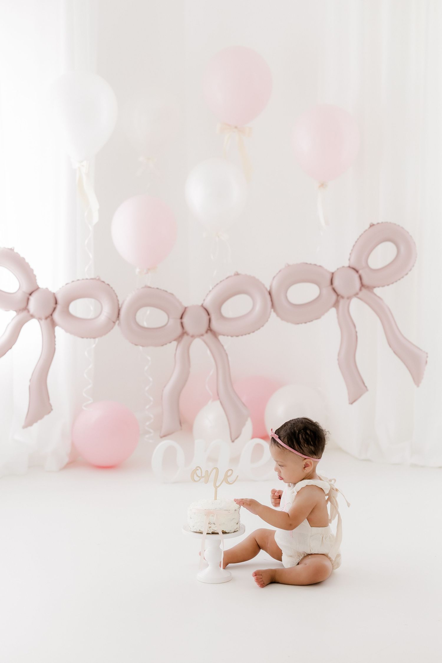 Sweet baby photography session with pink bows and white balloons in a dreamy studio setting.