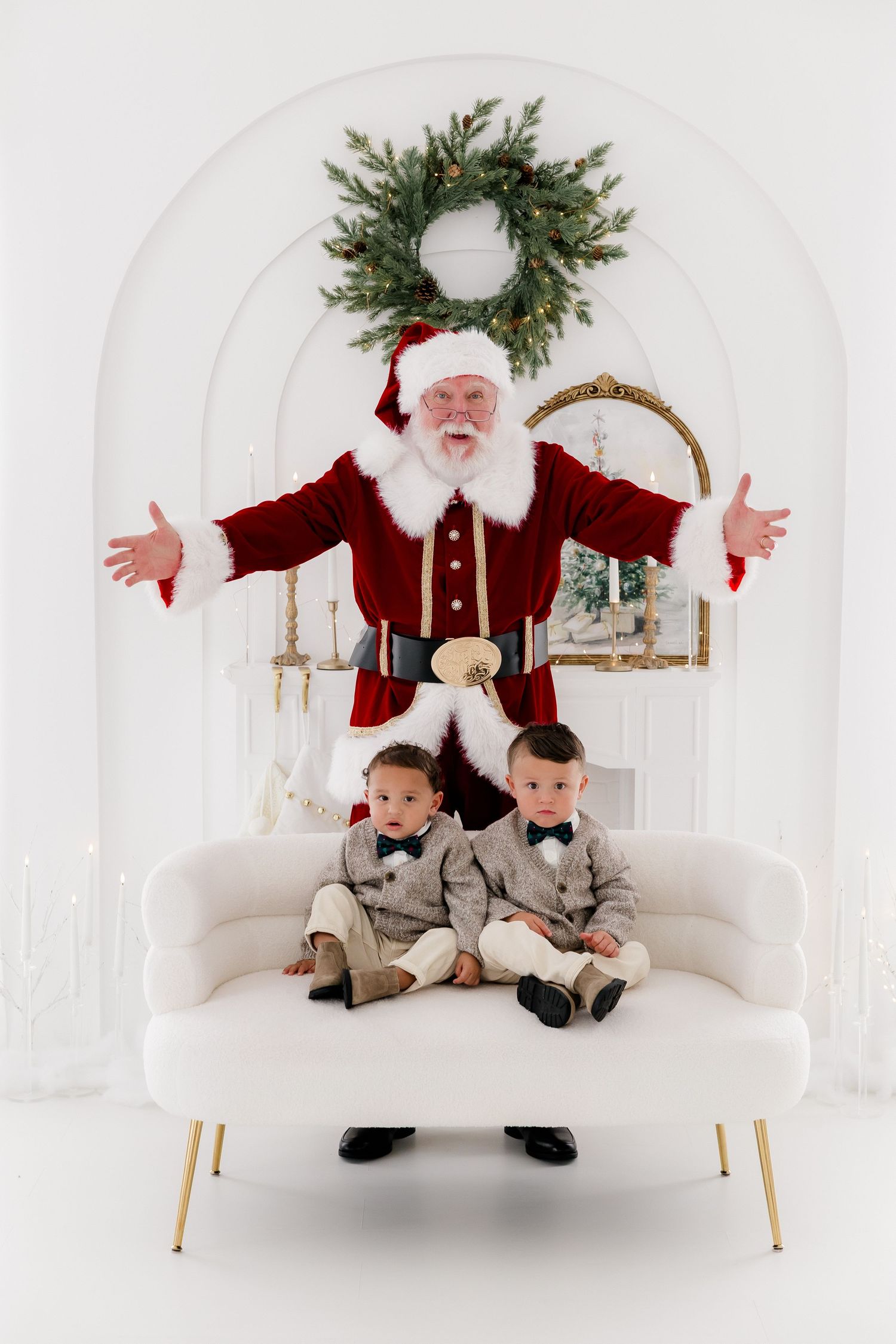 Two young boys in matching suits sit on a white sofa with Santa Claus behind them in a festive holiday photo.