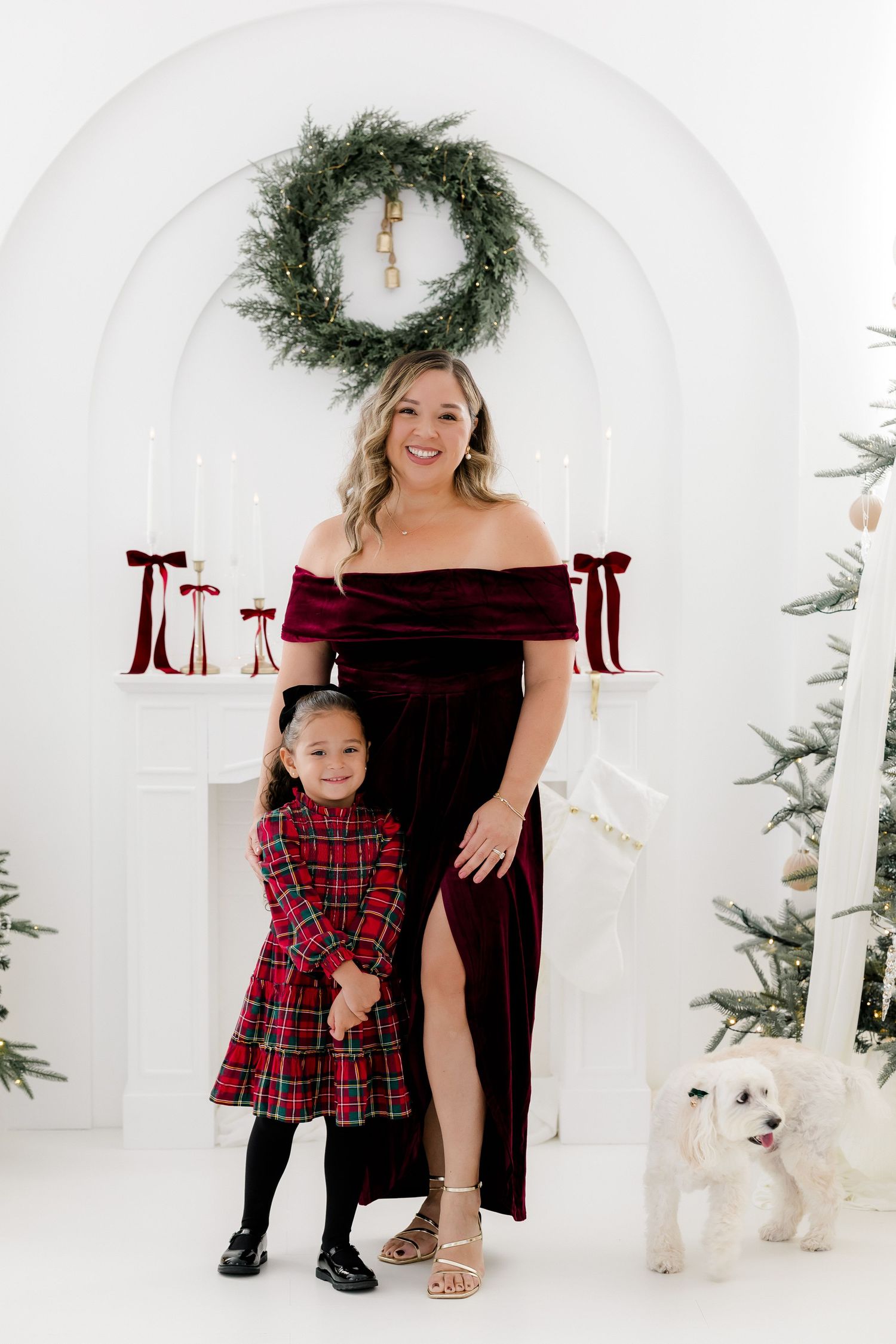 Woman in burgundy velvet gown poses with young girl in plaid dress and white dog before Christmas fireplace.