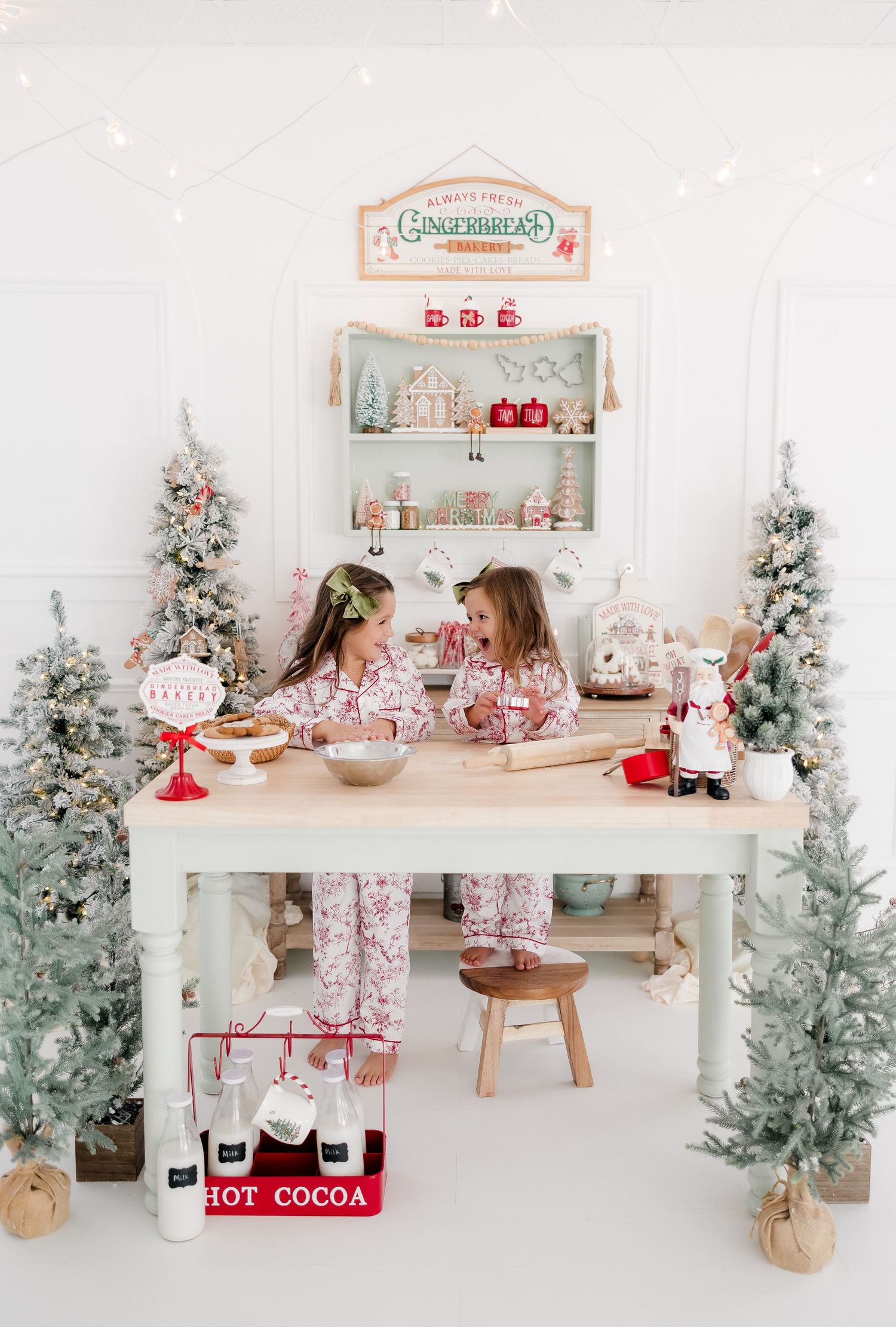 Two girls in matching pajamas bake Christmas cookies at a decorated table with flocked trees and hot cocoa in a festive white studio.