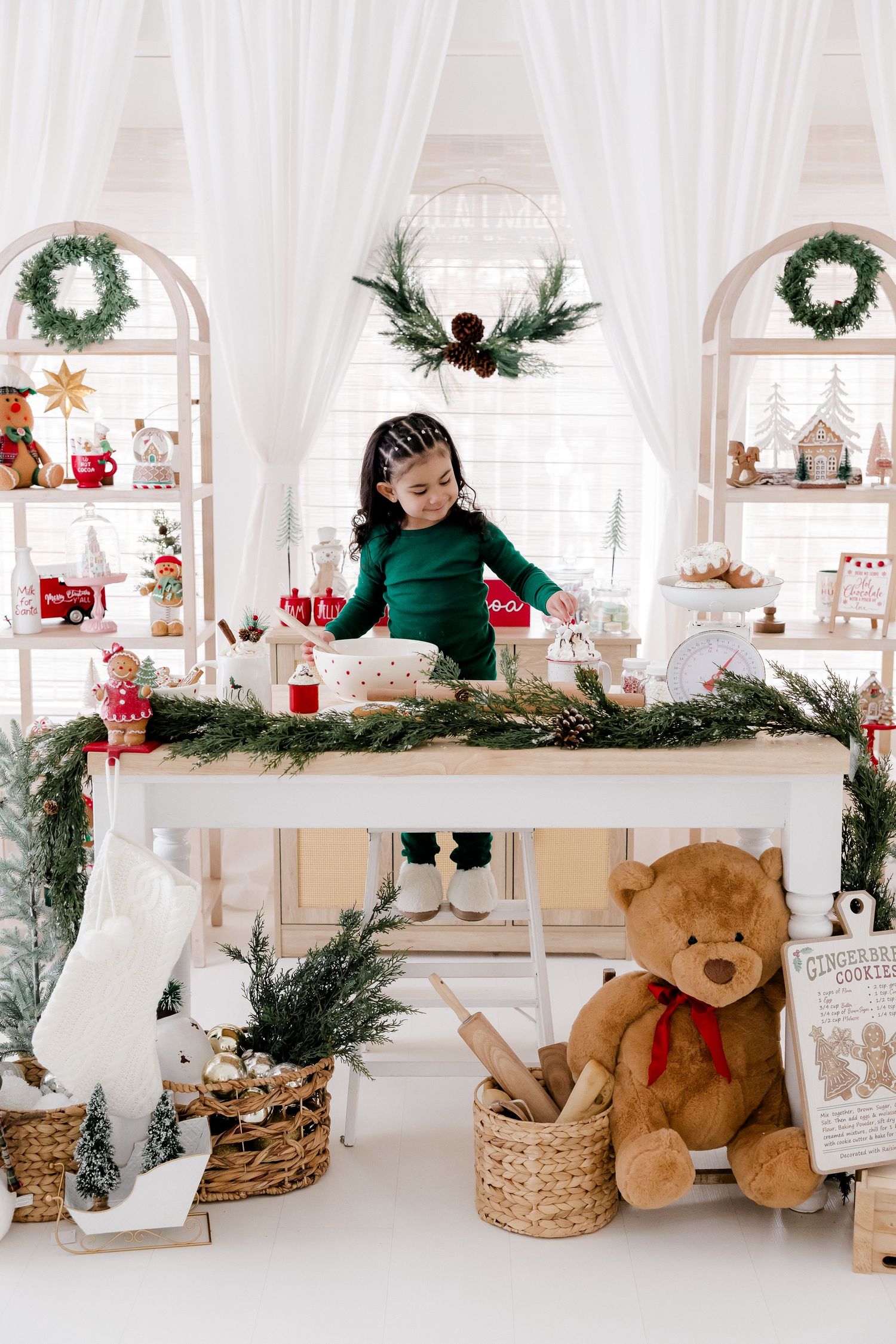 White holiday display setting with teddy bears, wreaths, and evergreen garland decorations.