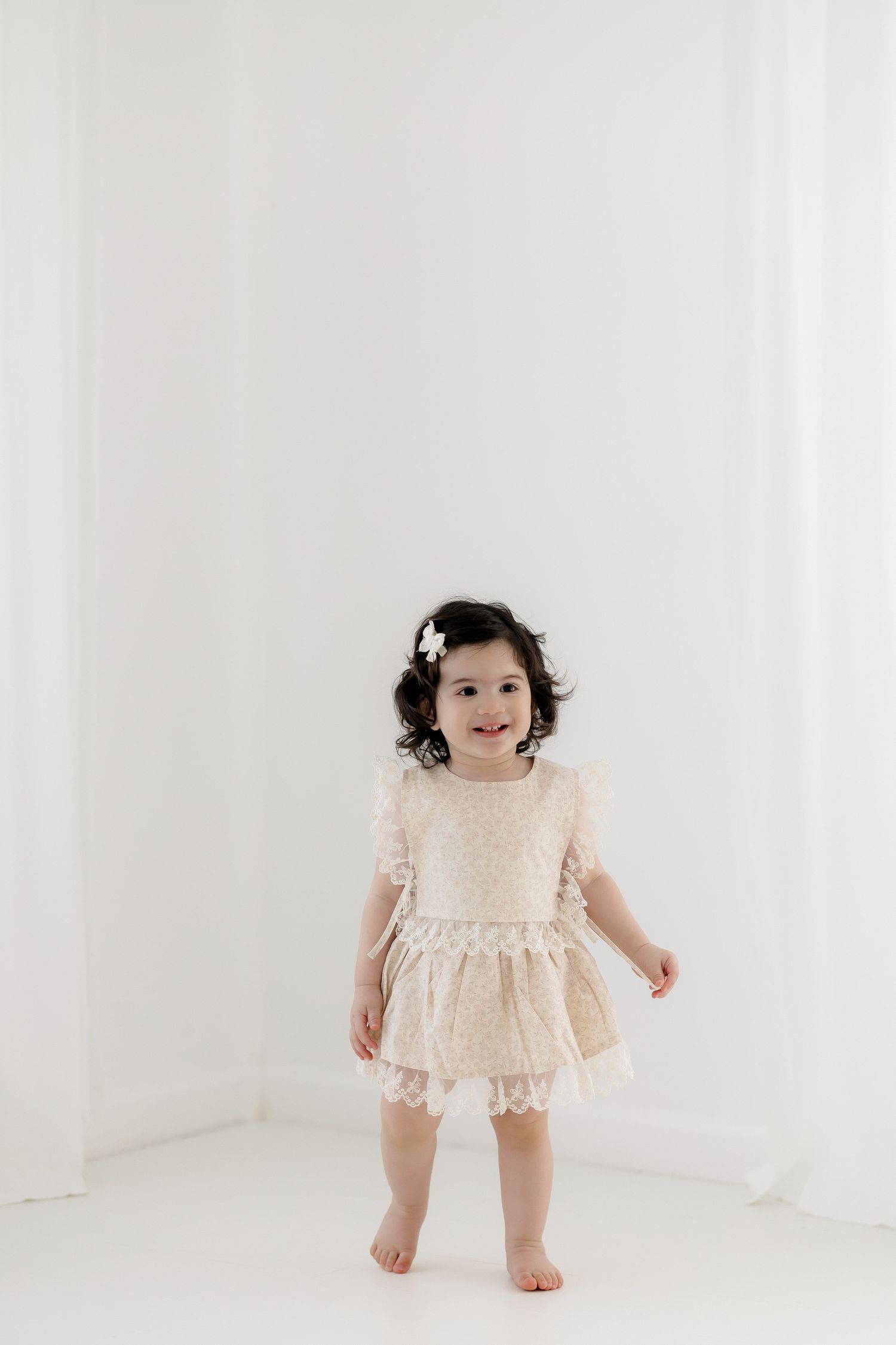 Smiling toddler girl in a cream lace dress with a flower hair clip stands barefoot in a bright white studio.