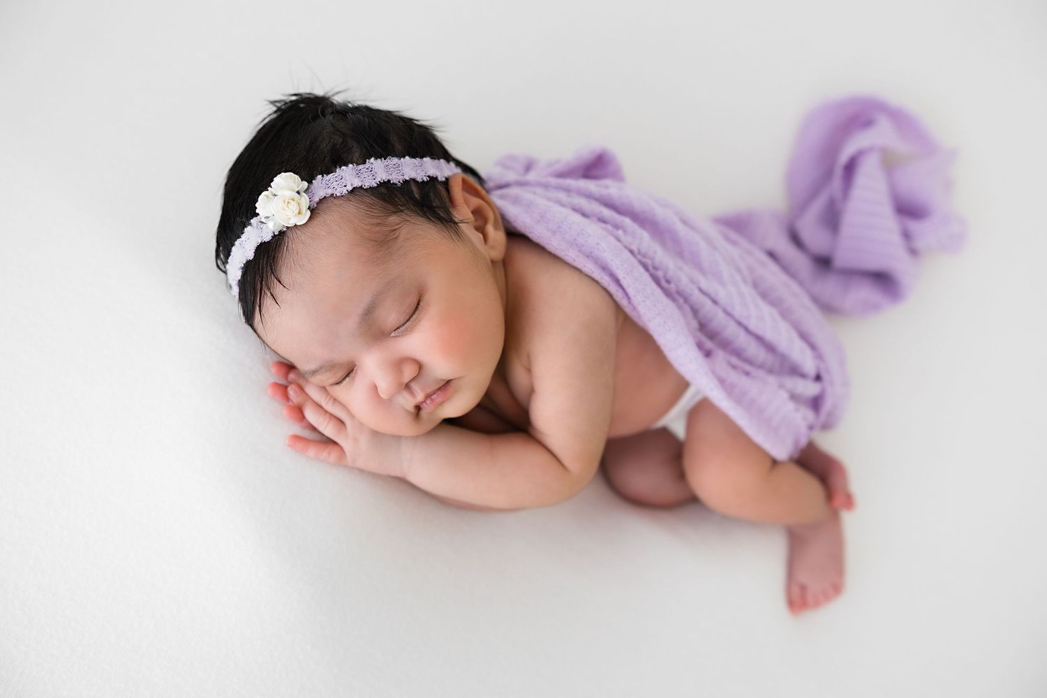 A sleeping newborn baby wrapped in soft purple fabric lies peacefully against a white backdrop.