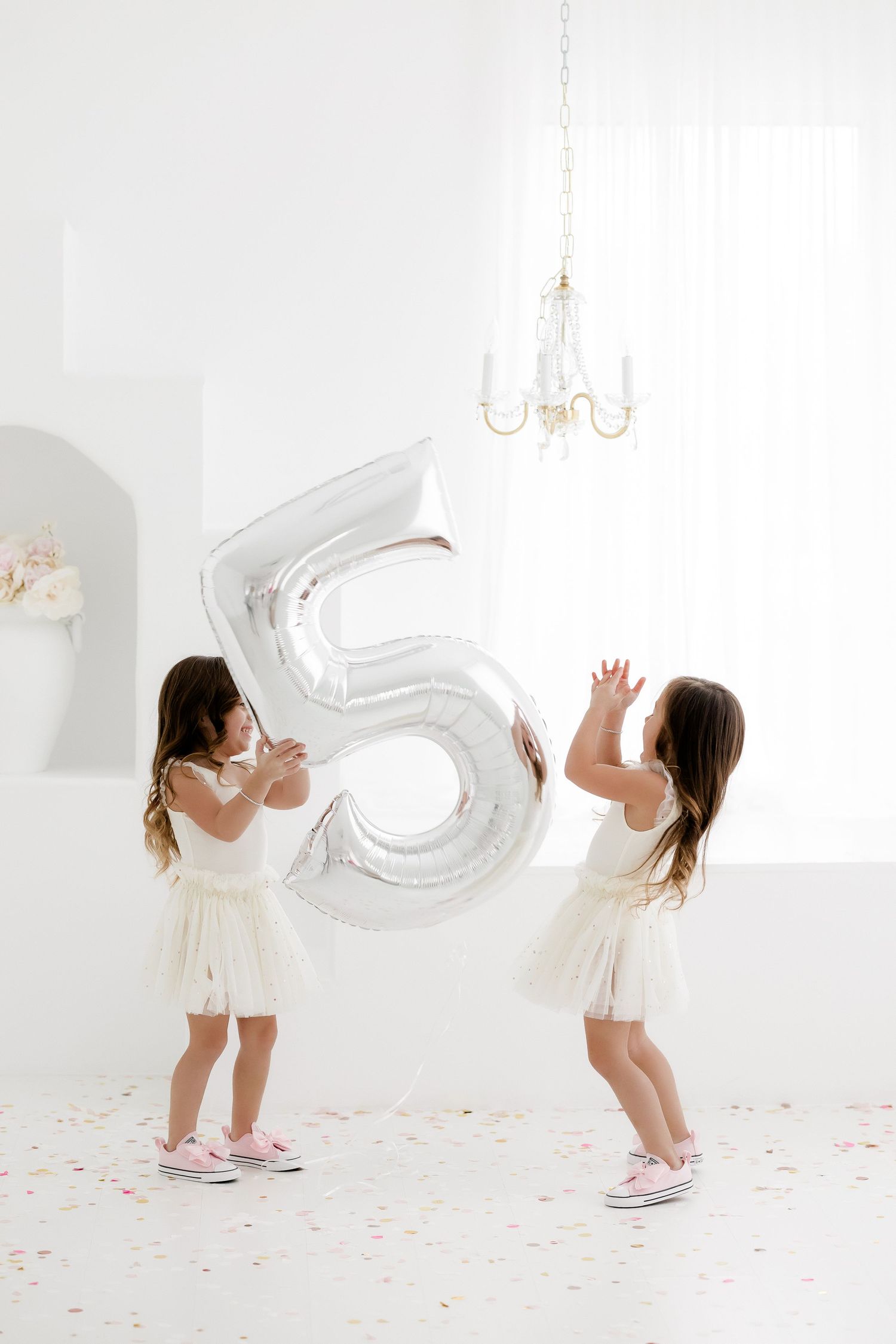 Child in white dress plays with silver number balloon in bright white studio setting with scattered confetti.