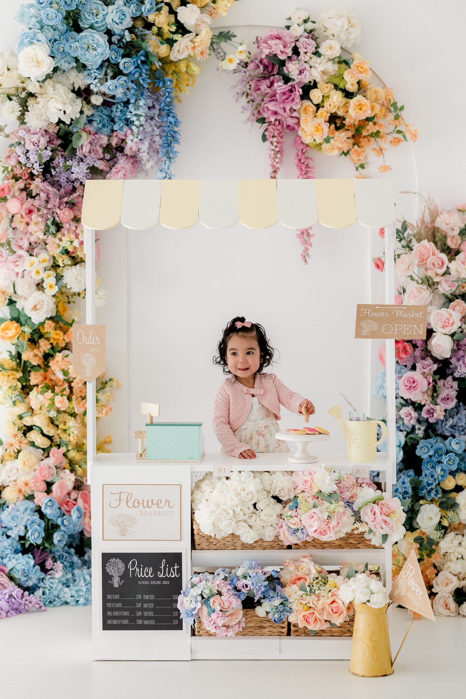 Whimsical flower stand display with pastel pink, blue, and yellow blooms surrounding a white booth with chalkboard menu.
