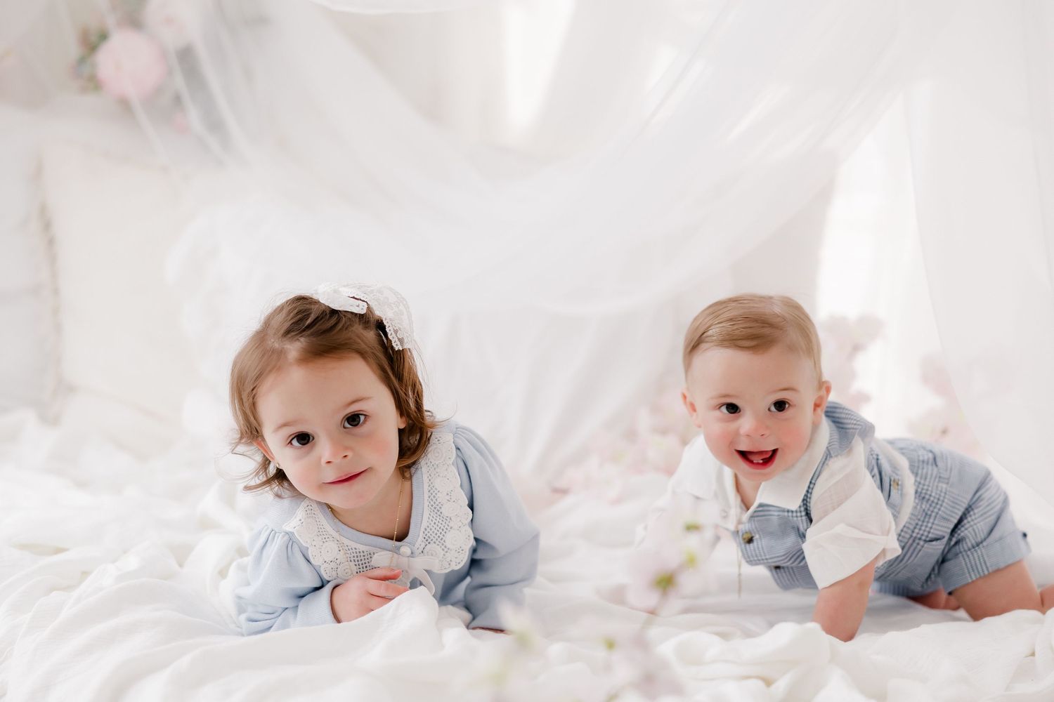 Two babies in light blue outfits lying on white bedding while smiling at the camera.
