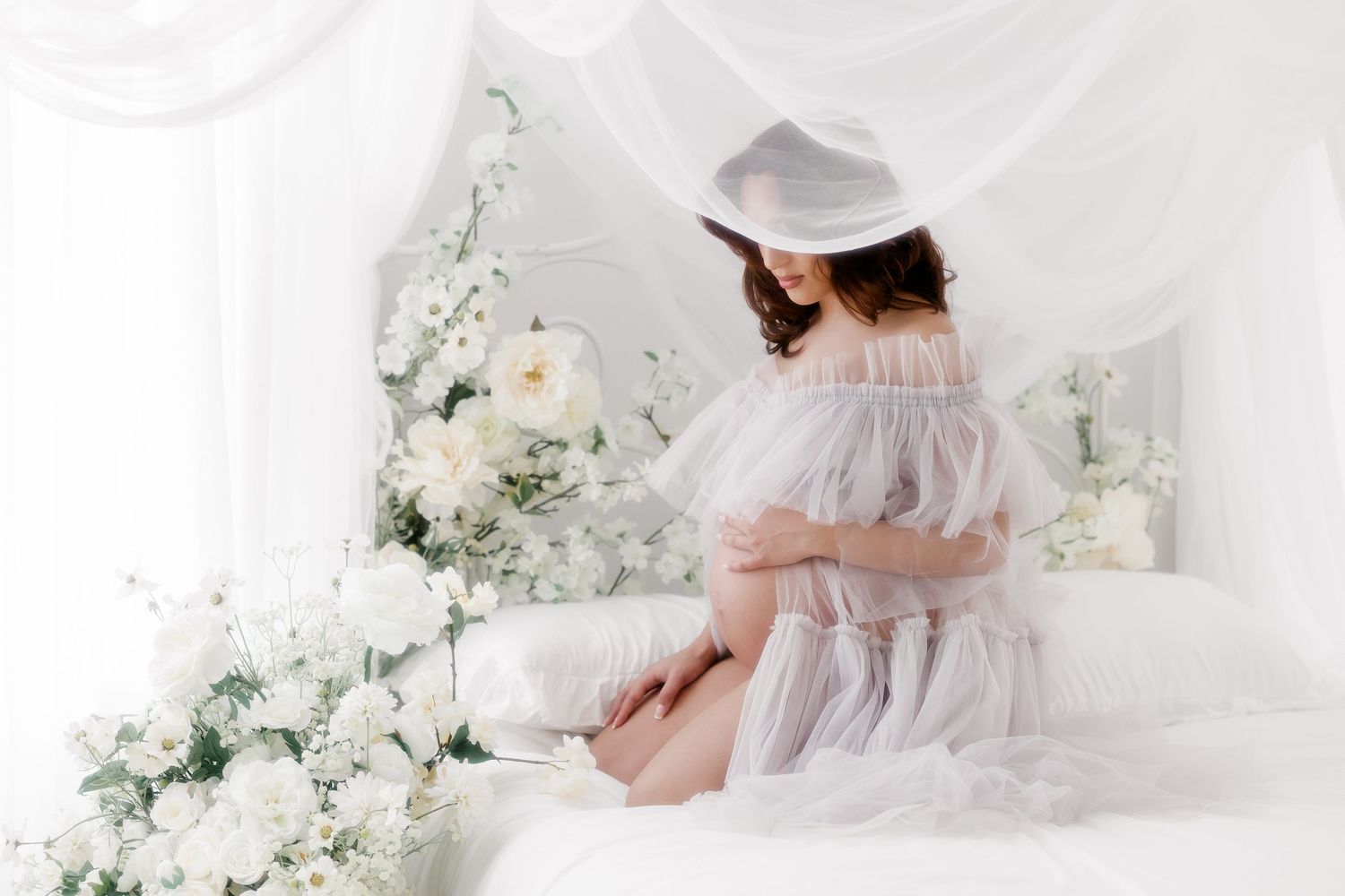 A delicate white lace and tulle maternity dress is showcased against a light, ethereal backdrop with floral decorations.