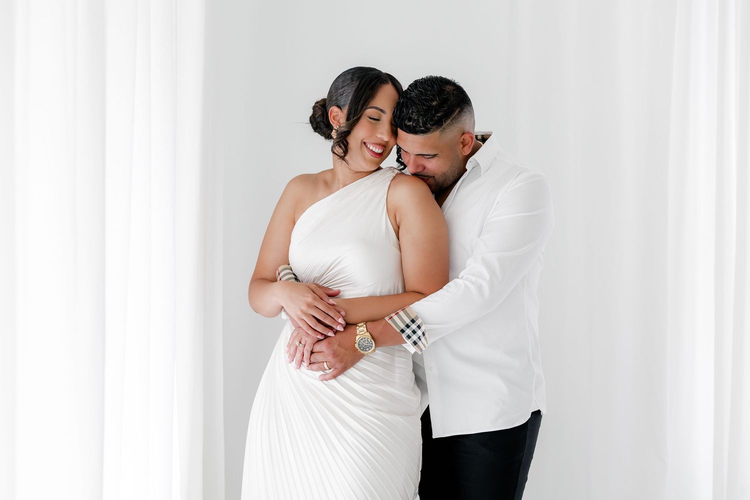 A romantic couple embraces in an intimate moment against a white backdrop, both wearing elegant white attire.