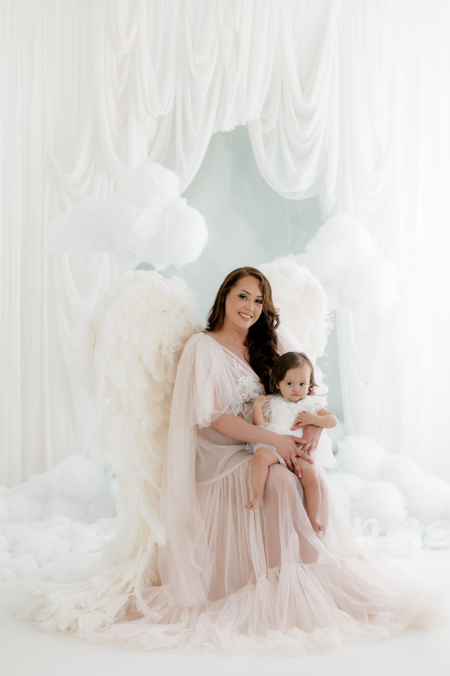 A mother in a white tulle gown with angel wings holds her baby amid white balloons and draped curtains.