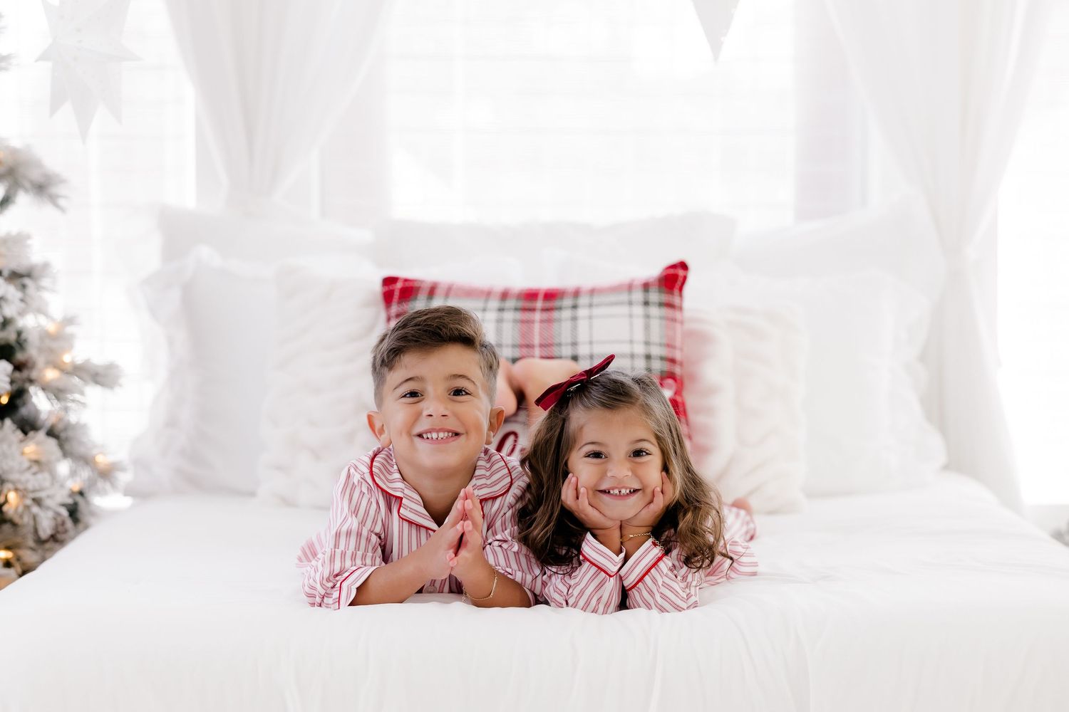 Two children in matching red striped Christmas pajamas lying on a white bed with a plaid pillow and holiday tree.