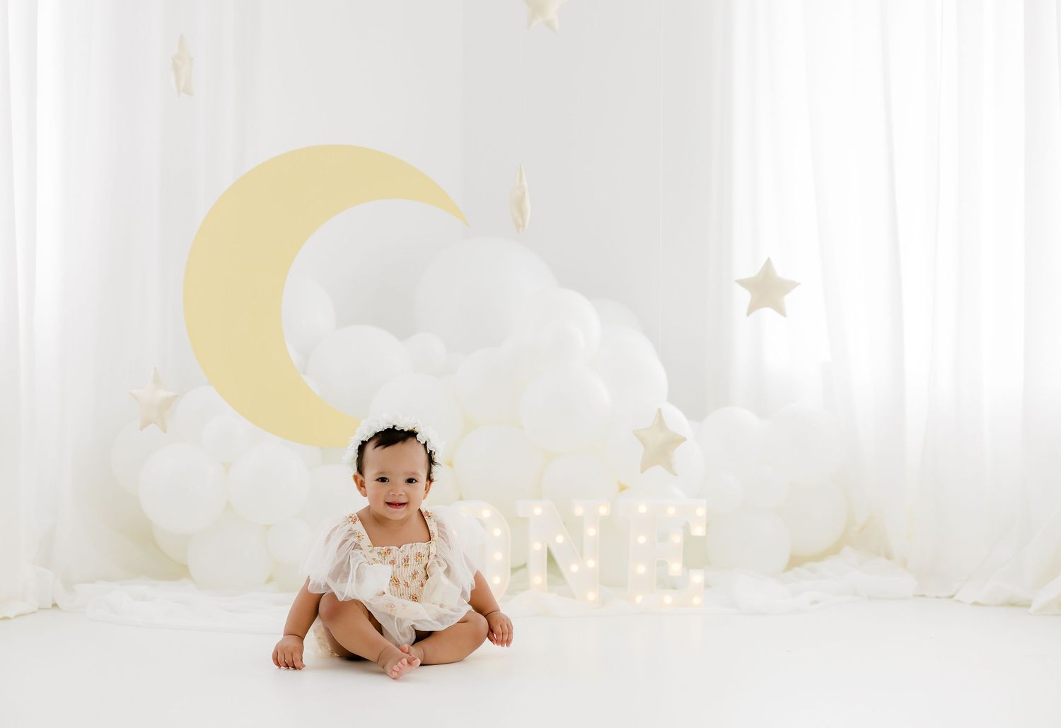 Baby sits on white floor with dreamy moon and cloud backdrop for nursery photography session.