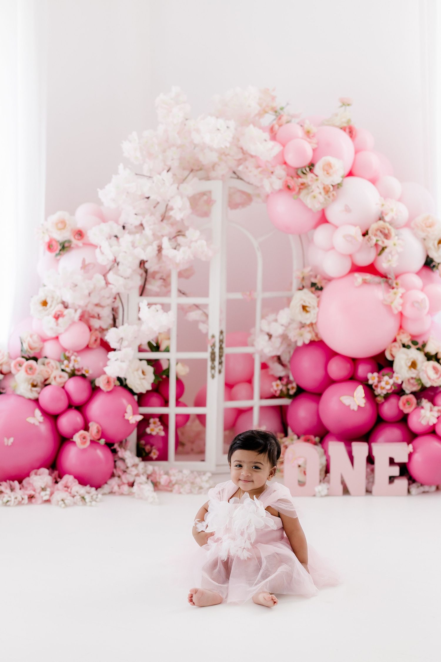 Pink balloon and floral backdrop with white window frame creates dreamy setup for first birthday photo session.