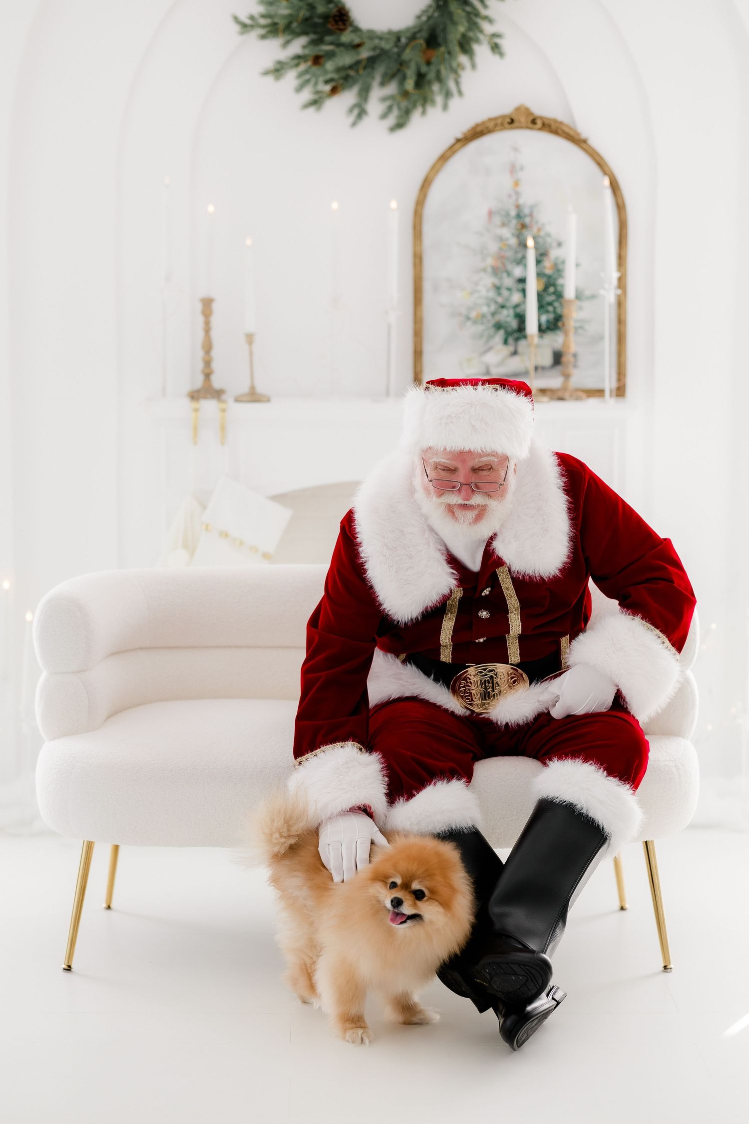 Santa Claus in red suit petting a fluffy Pomeranian dog in a white Christmas decorated room.