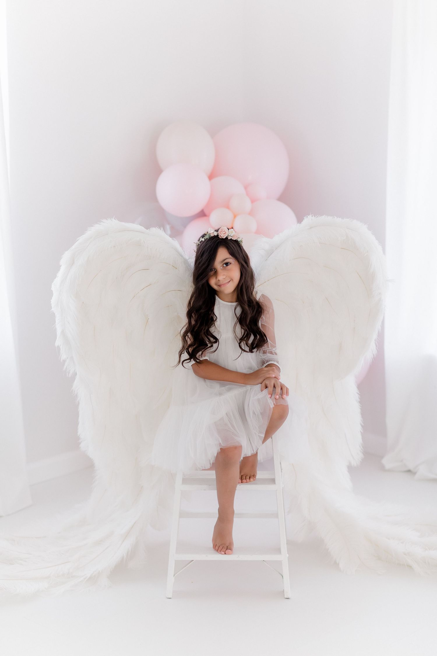 Angelic portrait featuring large white wings and pink balloons in dreamy studio setting.